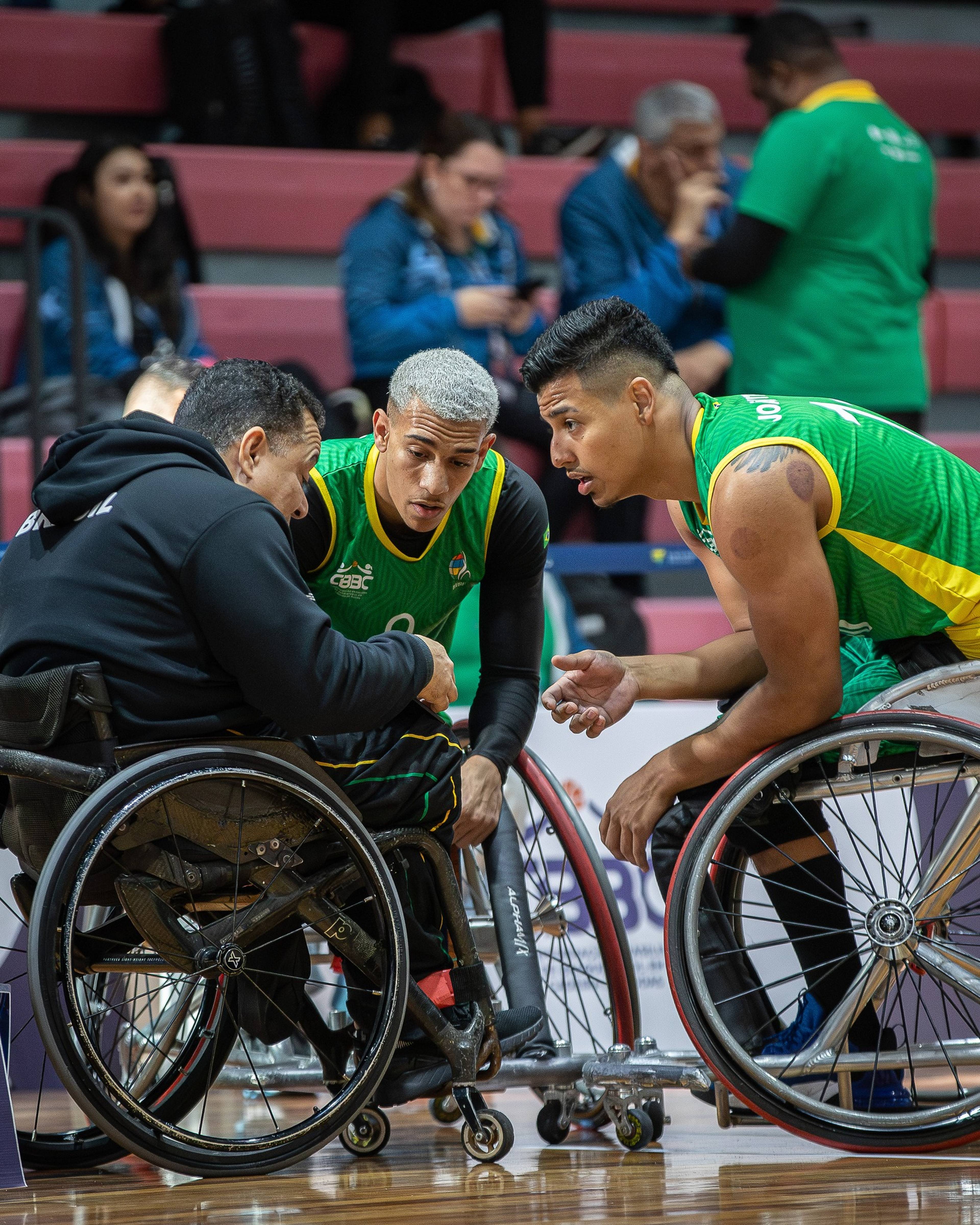 Two wheelchair basketball players in green jerseys listen intently to their coach during a game, with spectators in the background.
