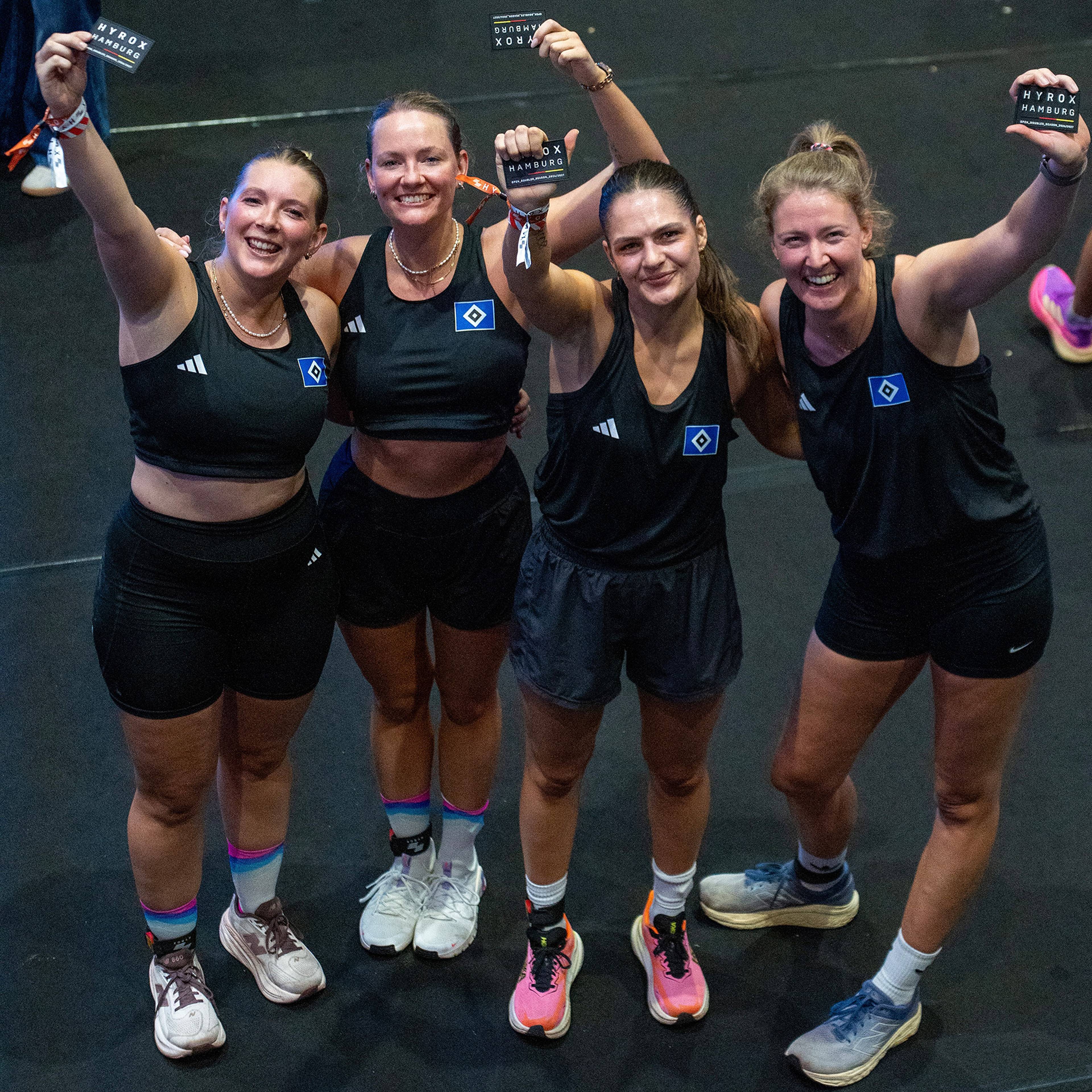 Four women in athletic attire smile and hold up cards, posing together in a gym setting.