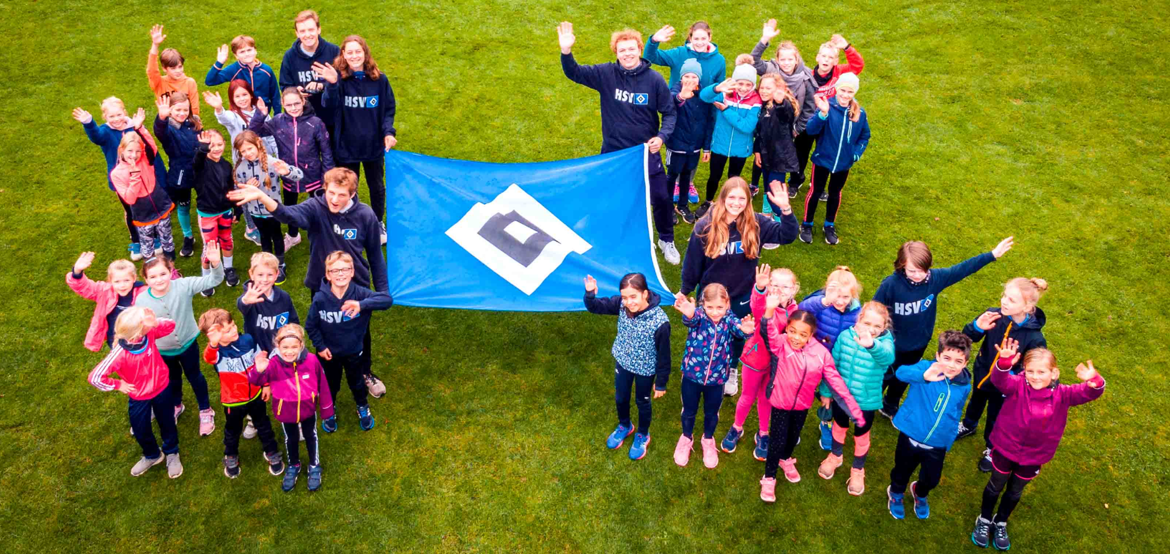 A group of children and adults wearing "HSV" hoodies wave on a grassy field, holding a blue flag with a black and white diamond logo.