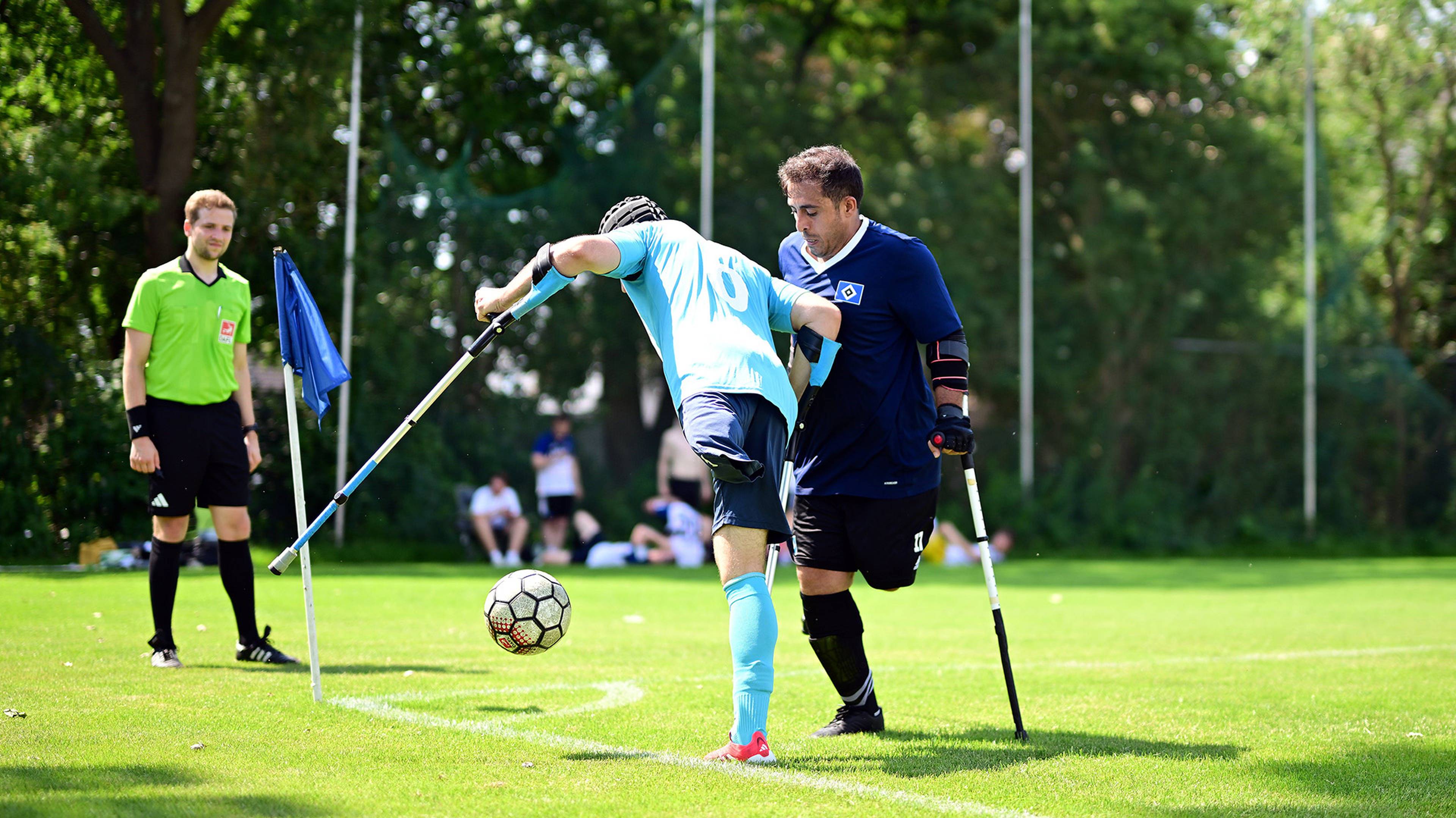 Two amputee soccer players compete on the field with crutches, observed by a referee in green.