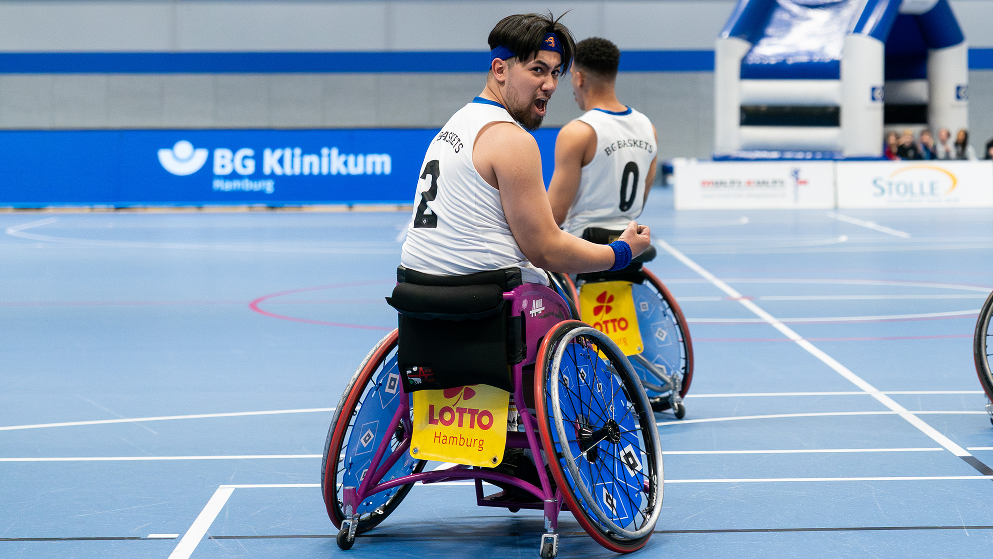 A wheelchair basketball player on the court passionately gestures with his arm, wearing a white jersey number 2 and a blue headband.