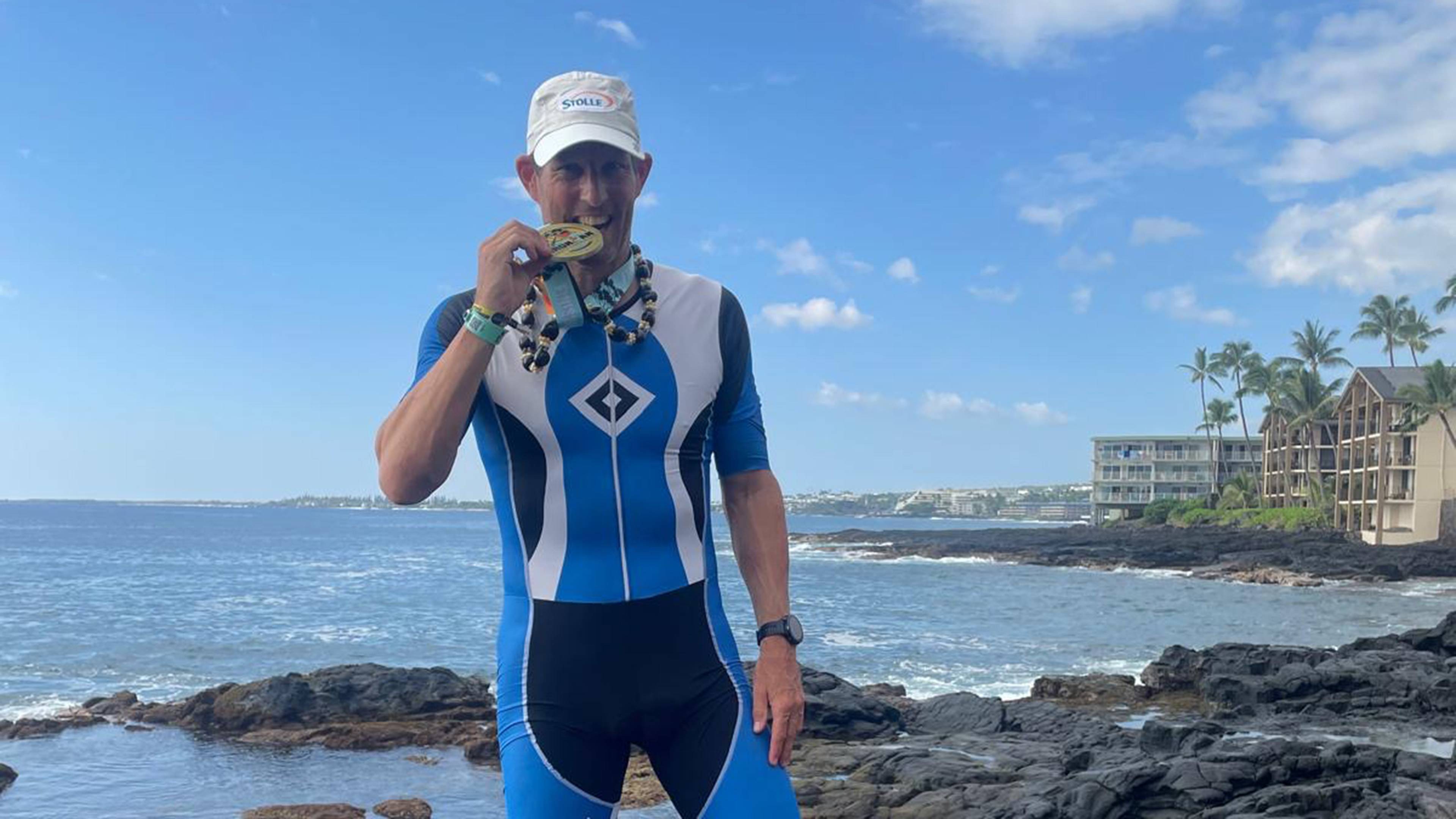 Man in a blue triathlon suit holding a medal to his mouth, standing on rocky shore with ocean and buildings in the background.