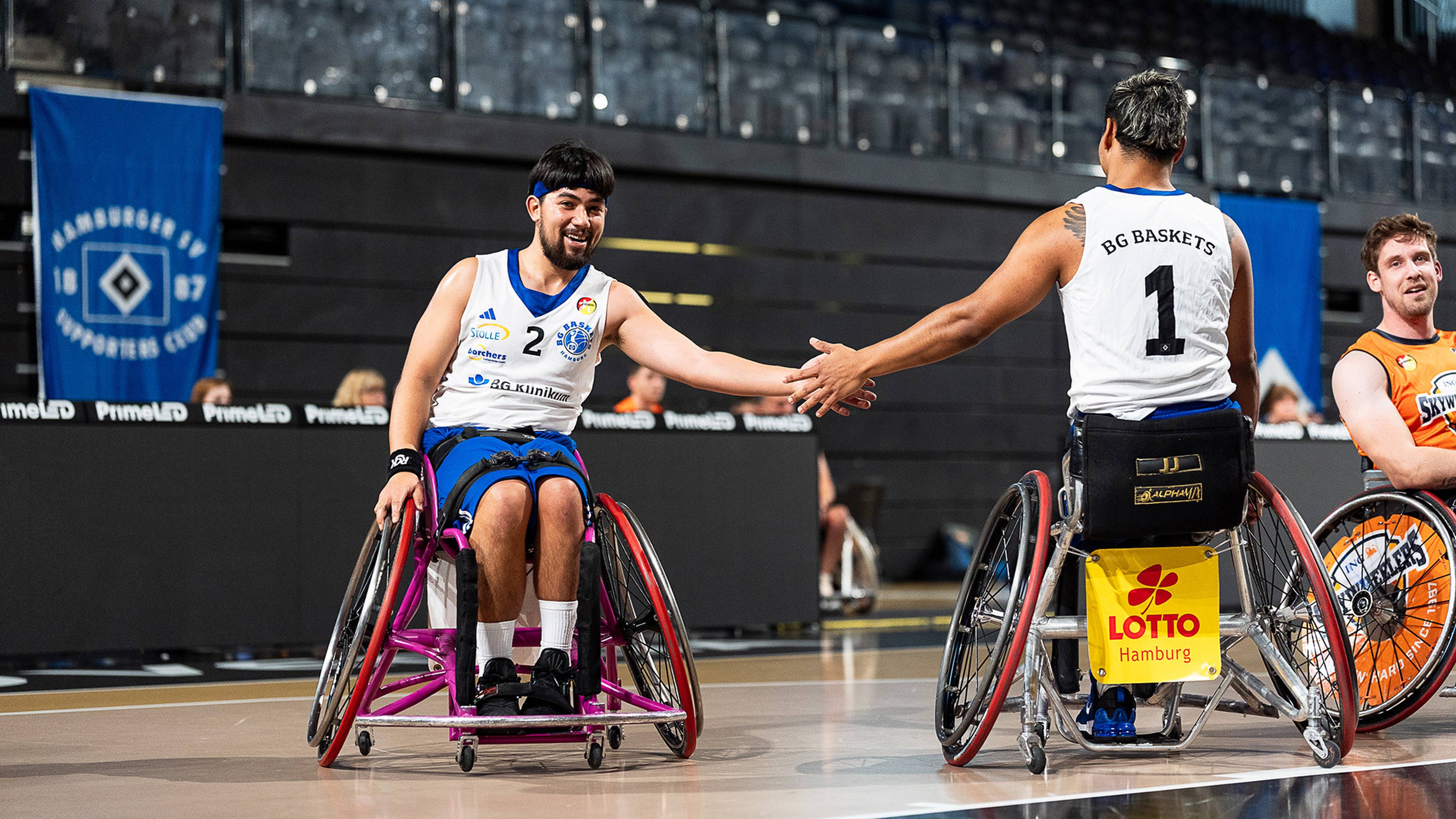 Two wheelchair basketball players smiling and high-fiving during a game, with spectators and banners in the background.