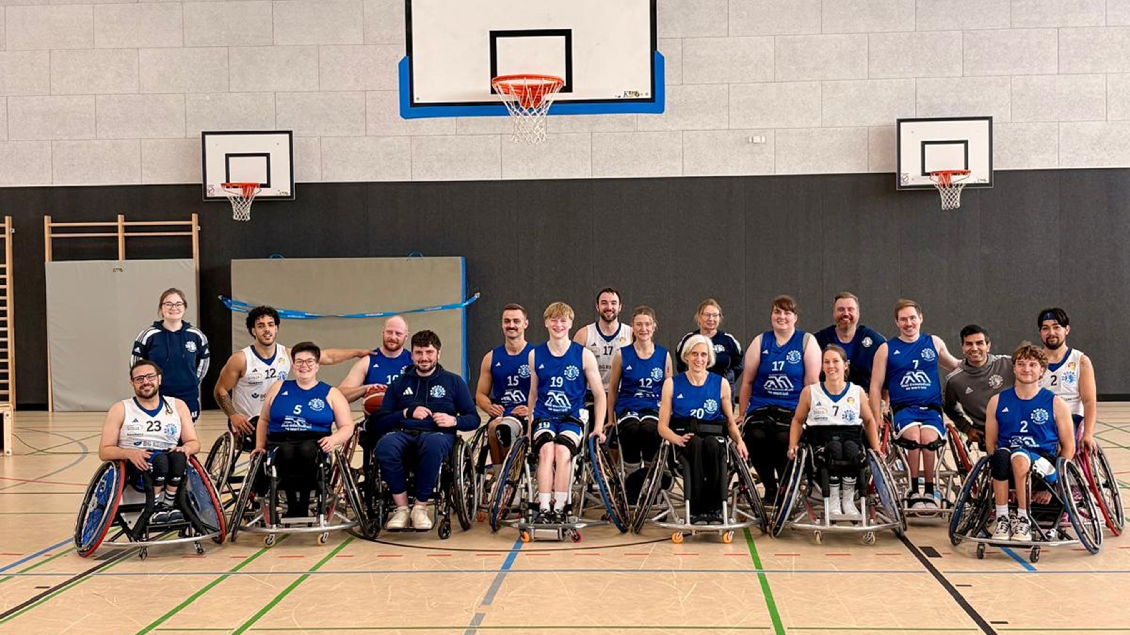 Group photo of a mixed-gender wheelchair basketball team in blue jerseys, posed in a gymnasium with two basketball hoops in the background.