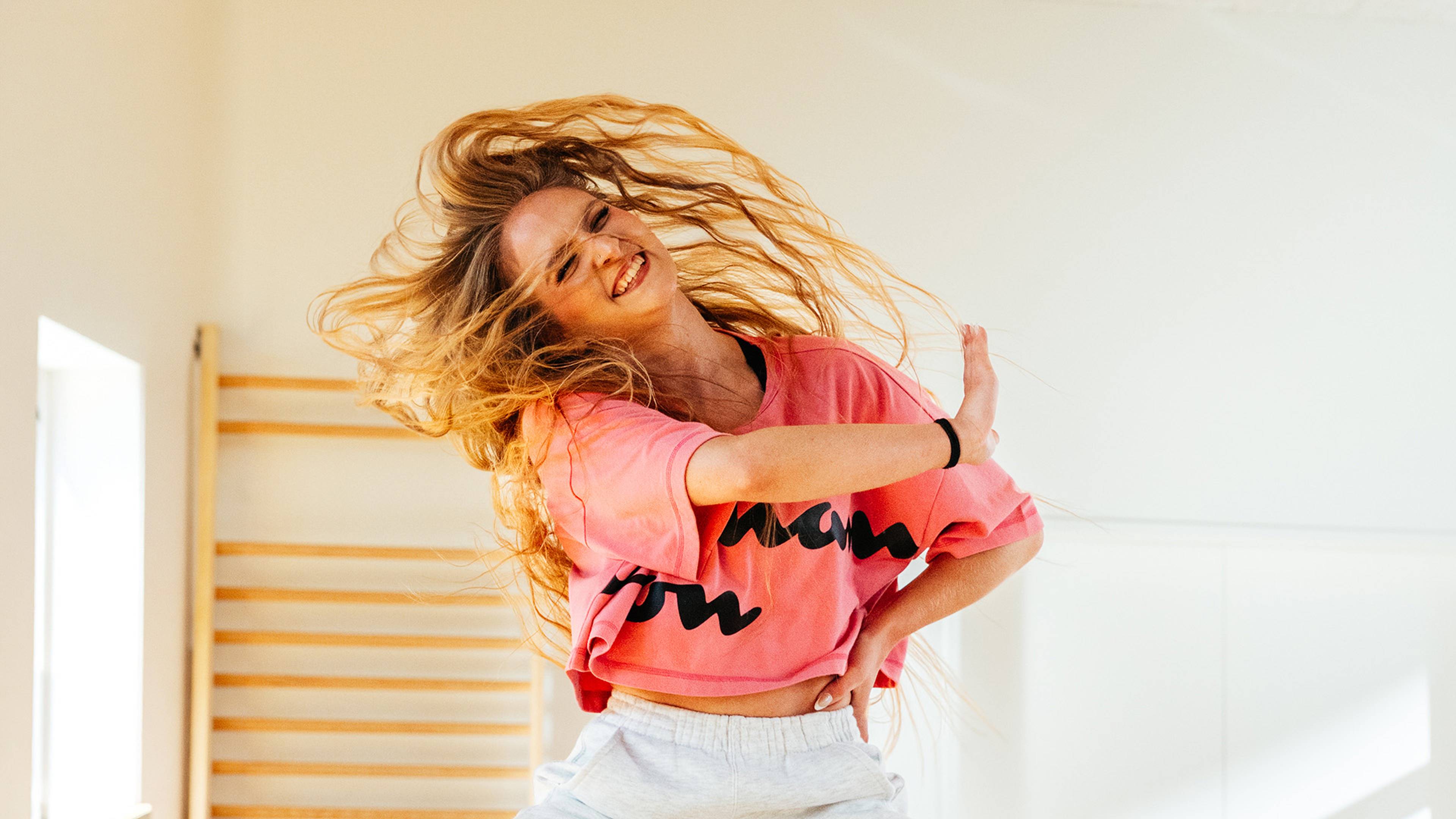 A joyful woman in a pink sweatshirt dances energetically, her long hair flowing, in a brightly lit room with wooden bars on the wall.