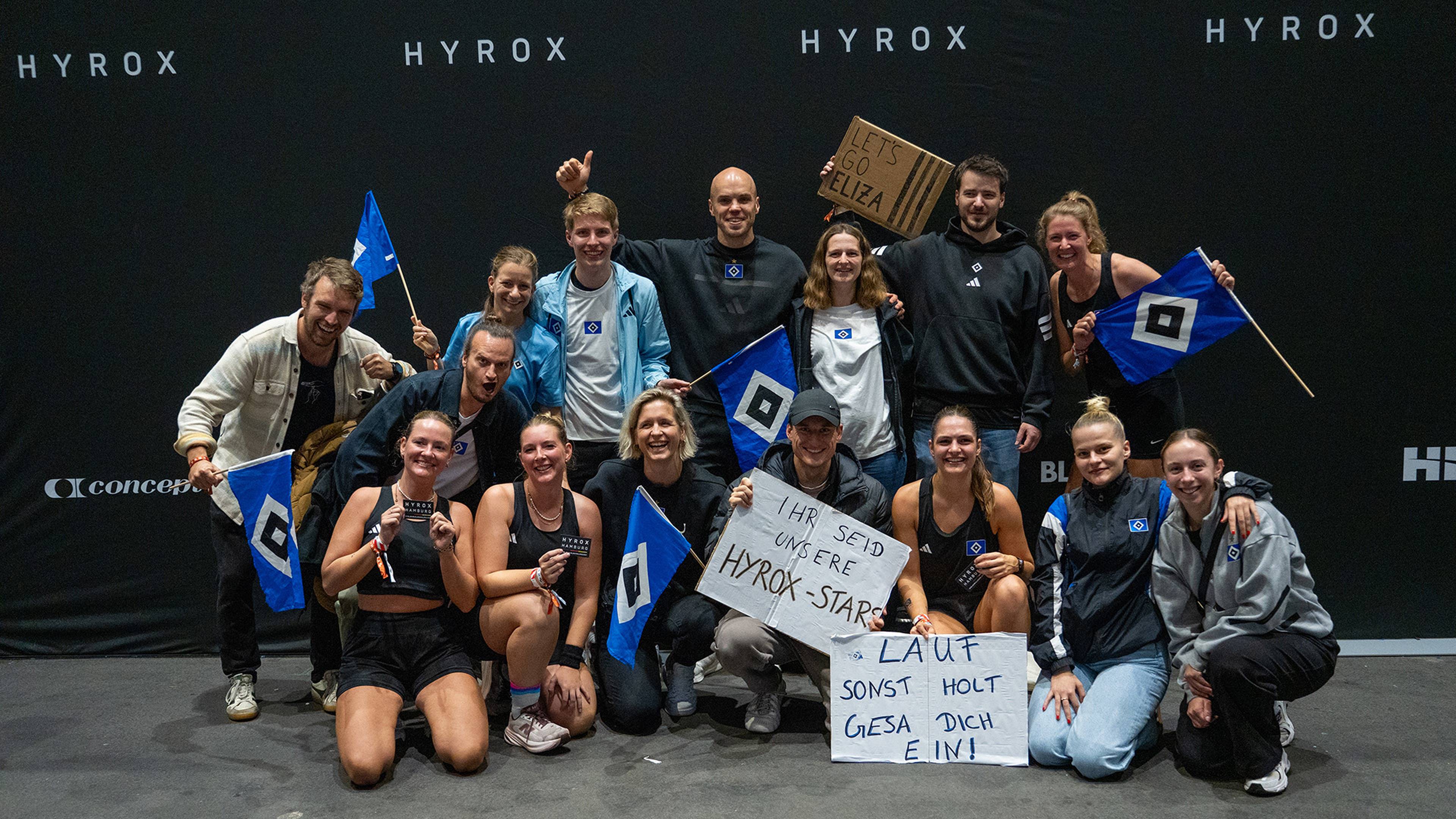 A group of people smiling and holding signs and flags at a Hyrox event, standing together in front of a black backdrop with "HYROX" text.