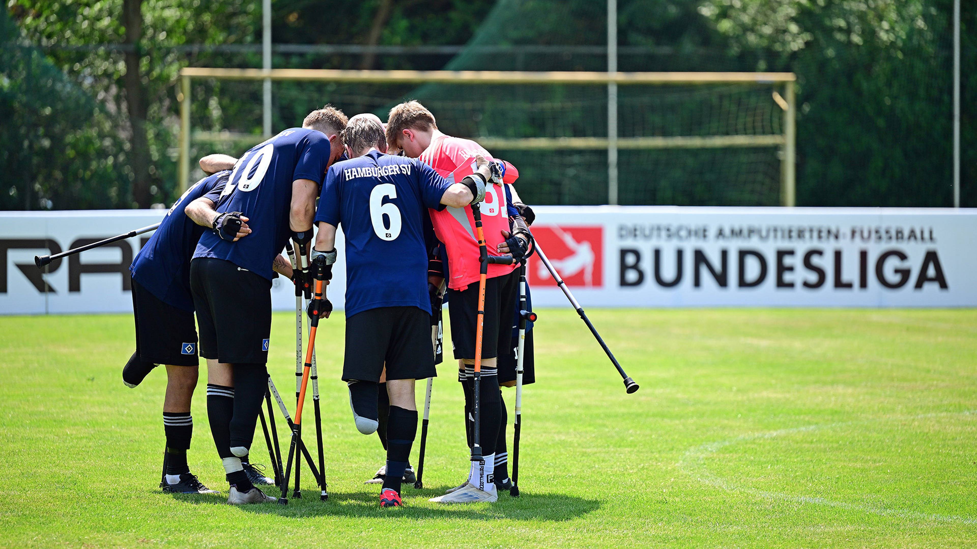 A group of amputee football players in a huddle on a grassy field, with a sign in the background reading "Deutsche Amputierten Bundesliga."