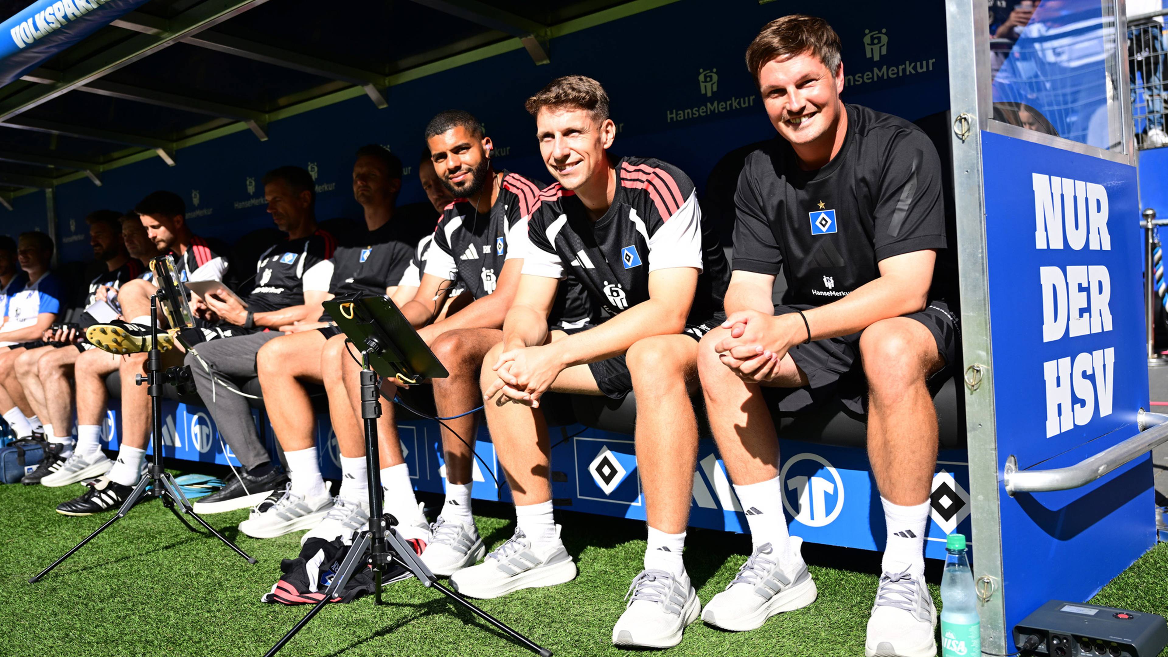 Cheftrainer Merlin Polzin und seine Trainerkollegen sitzen lächelnd auf der Trainerbank