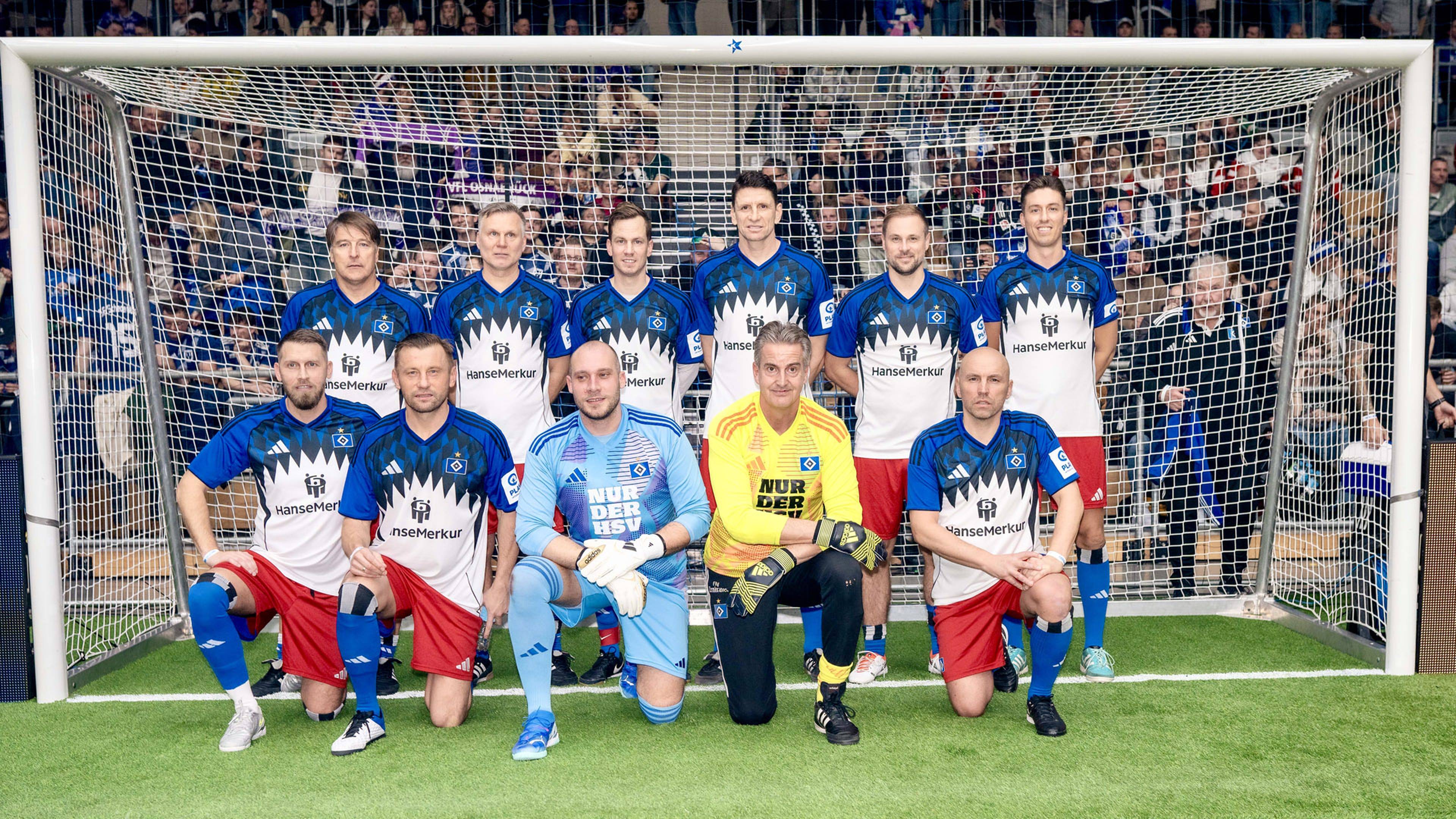 Eine Fußballmannschaft, bestehend aus elf Männern steht in einem Fußballtor hintereinander aufgestellt. Neun von ihnen tragen Feldspielertrikots, zwei Torwartkleidung. 