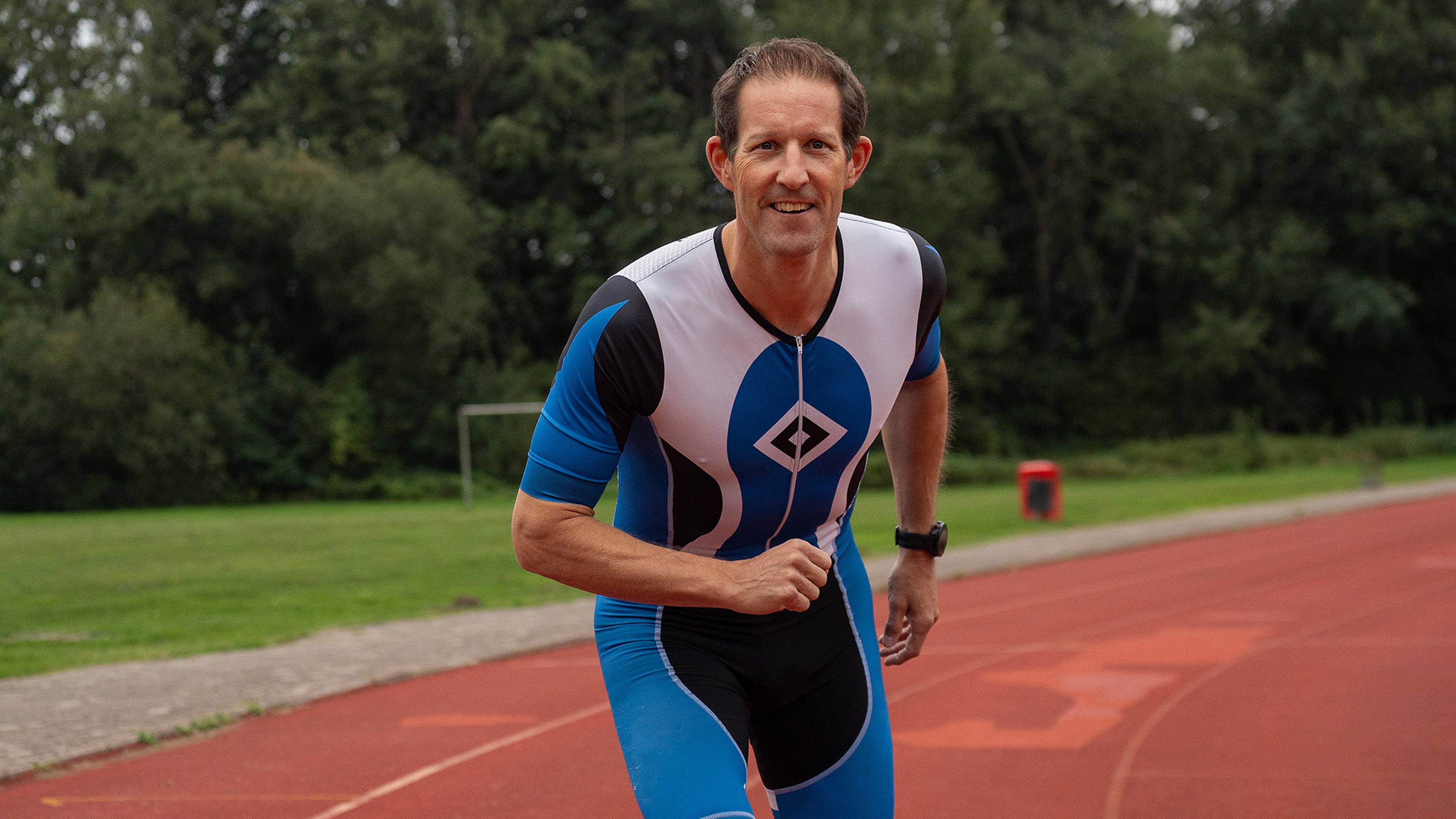 A man in a blue and white sports suit running on a track, surrounded by greenery, smiling and looking at the camera.