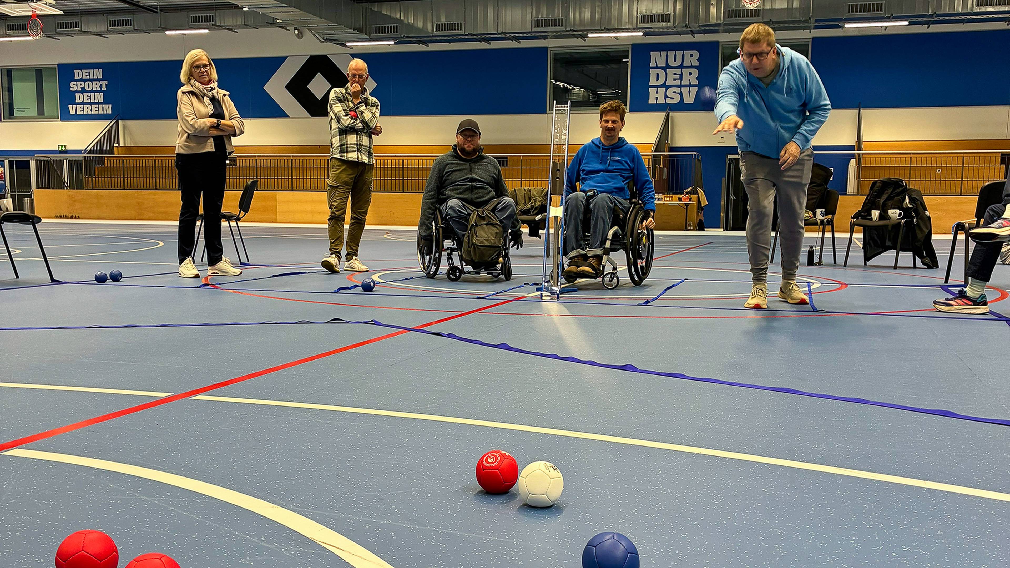 People in a gym playing boccia, with two participants in wheelchairs and others watching, blue and red balls scattered on the floor.