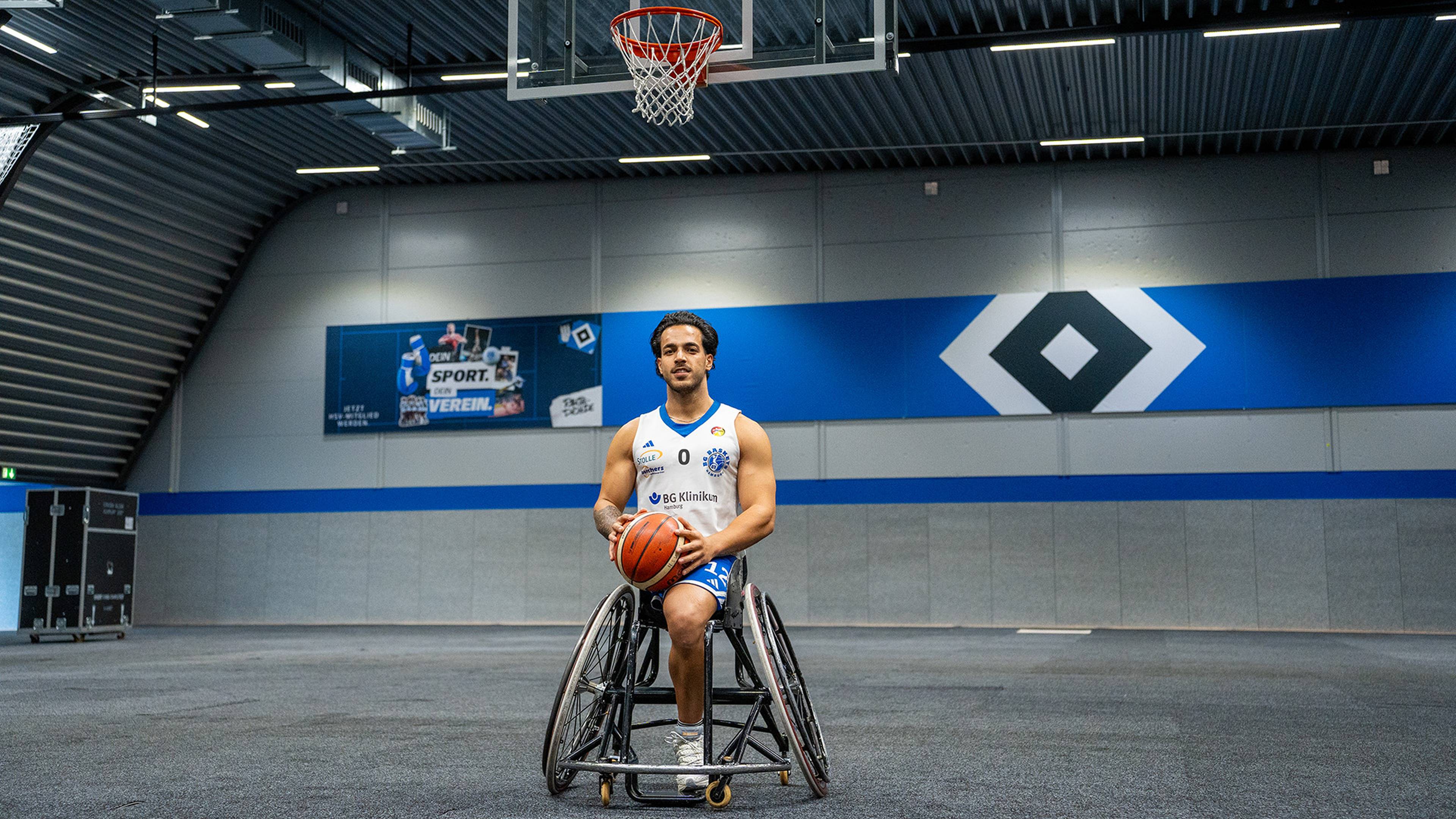 A basketball player in a wheelchair holds a ball on an indoor court, with a basketball hoop overhead and a blue logo on the wall behind.