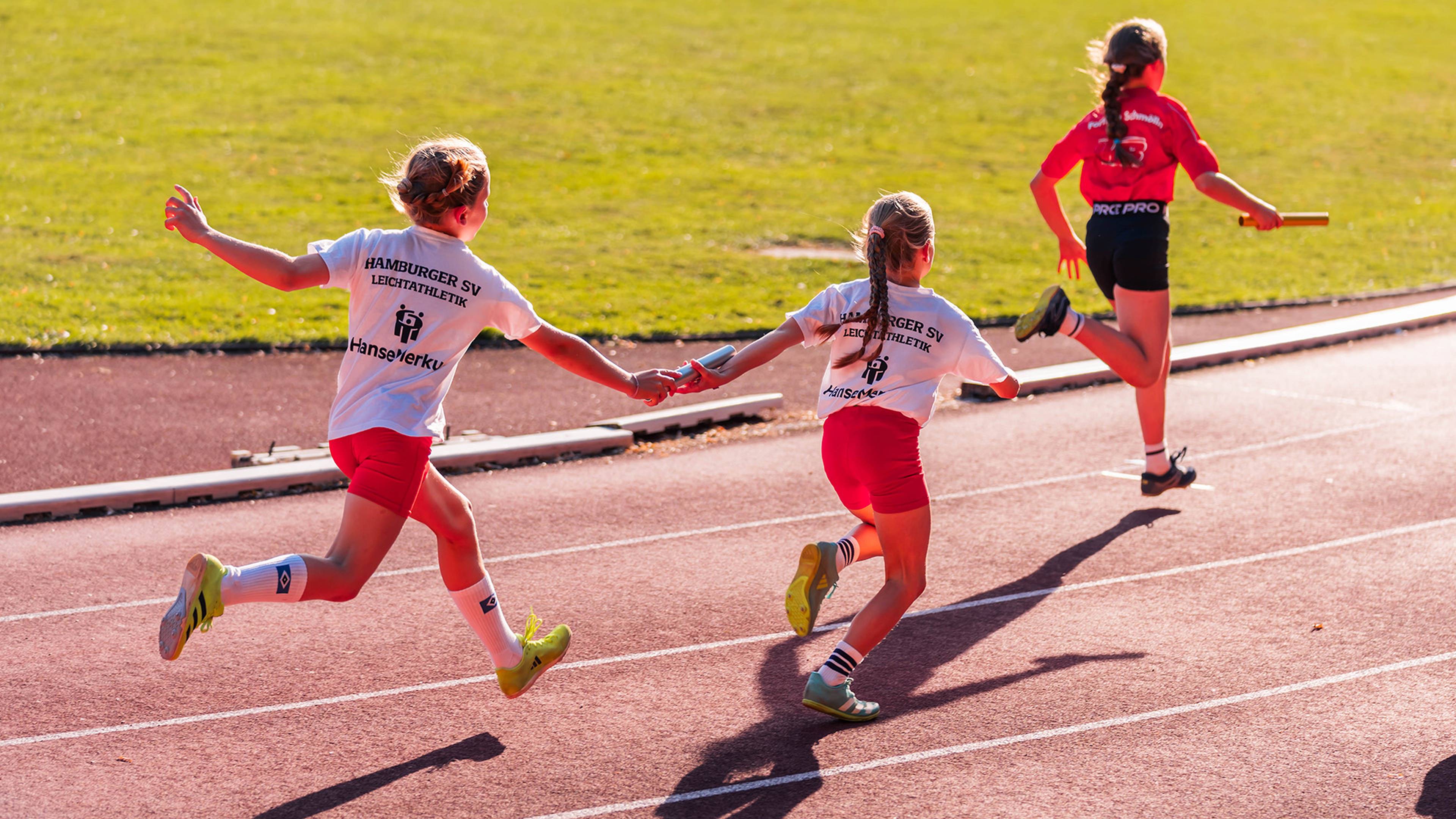 Children in athletic gear participate in a relay race on a track, with one handing a baton to another.