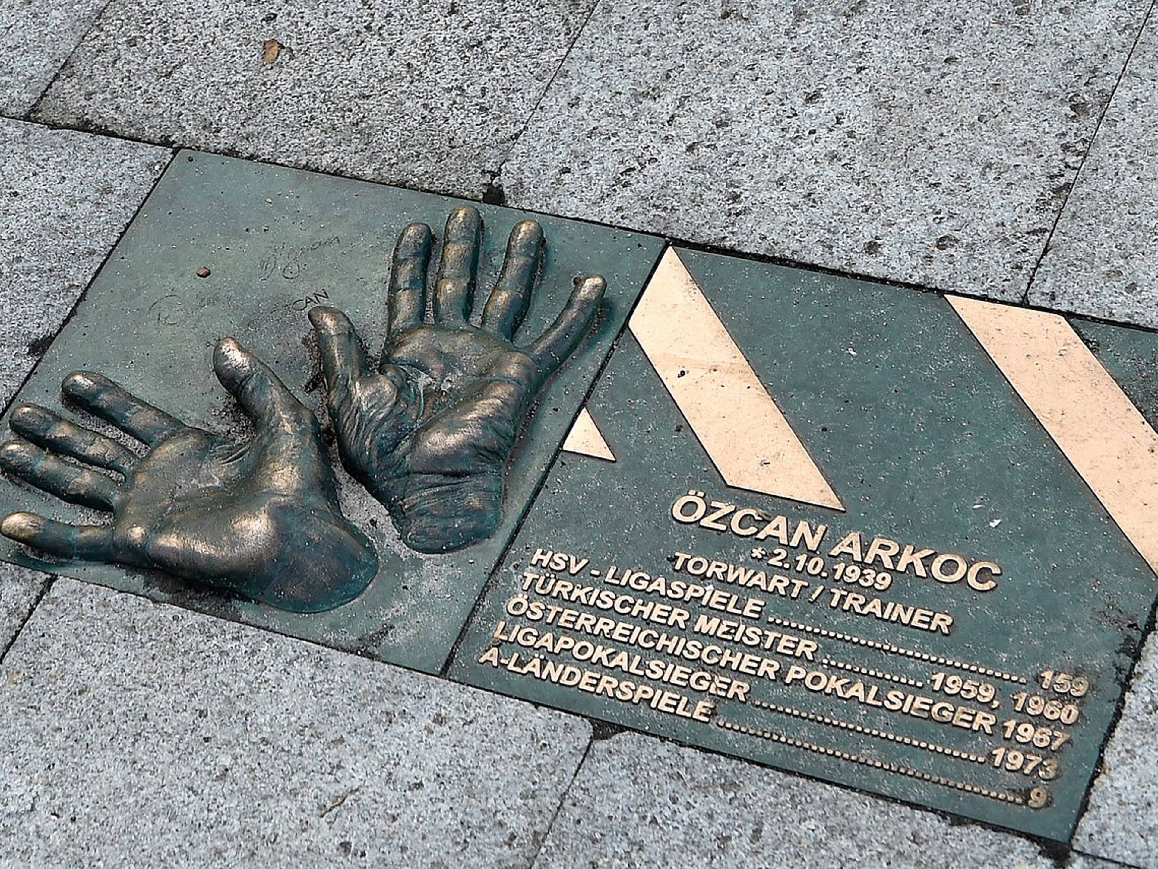 Bronzene Handabdrücke und eine Gedenktafel zu Ehren von Özcan Arkoc mit Informationen über seine Erfolge als Fußballtorwart und -trainer, eingelassen in einen Pflasterstein.