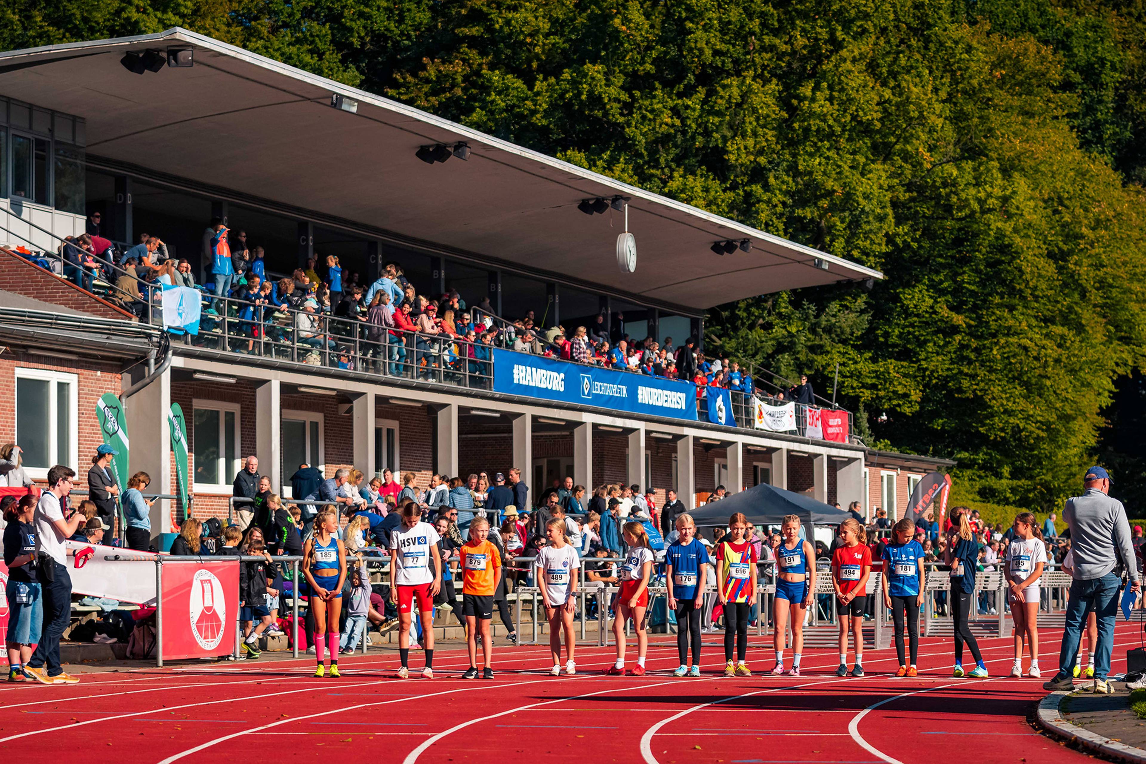 A group of young athletes stand on a track, preparing to race. Spectators fill the stadium seats, with trees in the background.