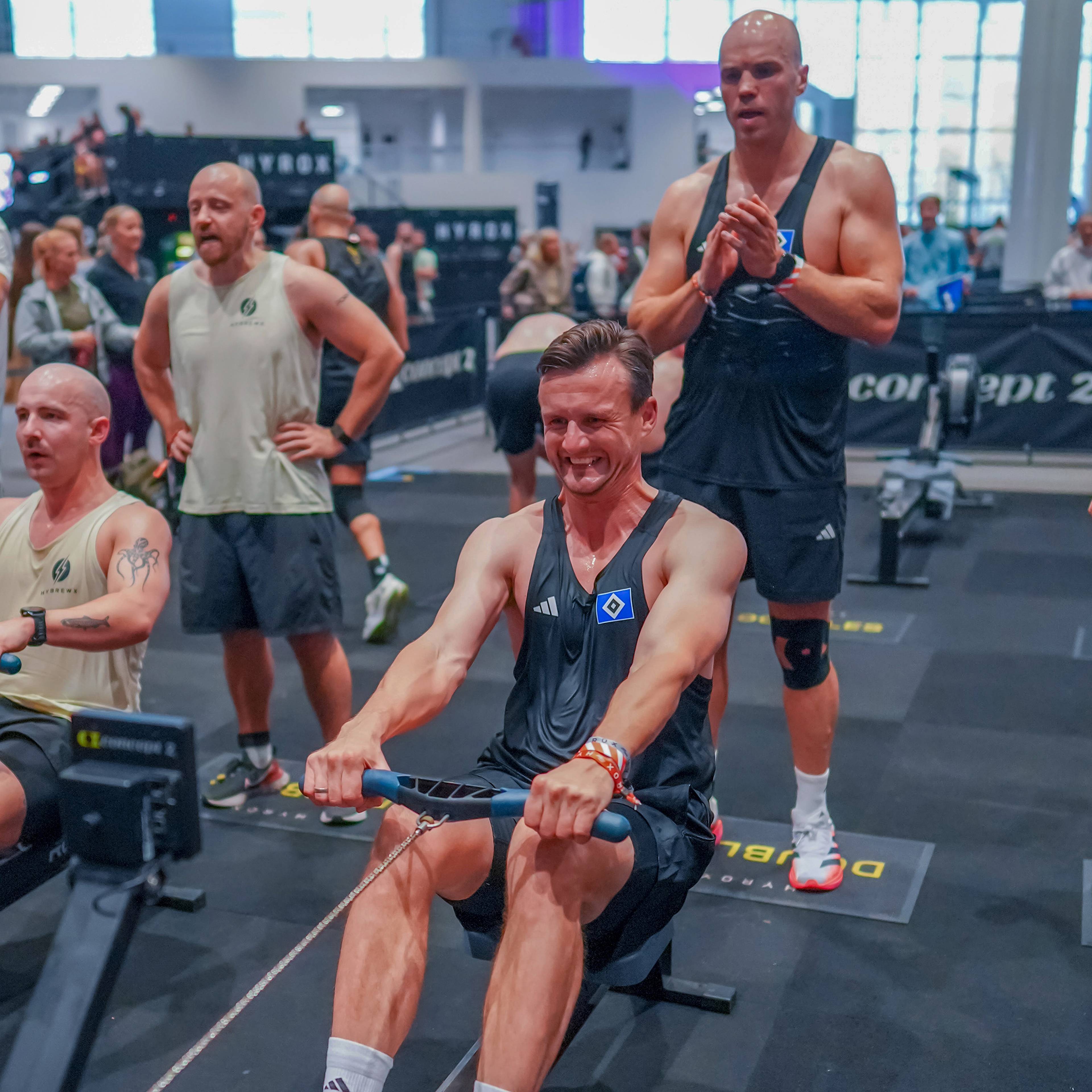 A man in athletic gear exerts effort on a rowing machine in a gym event setting, surrounded by spectators and other participants.