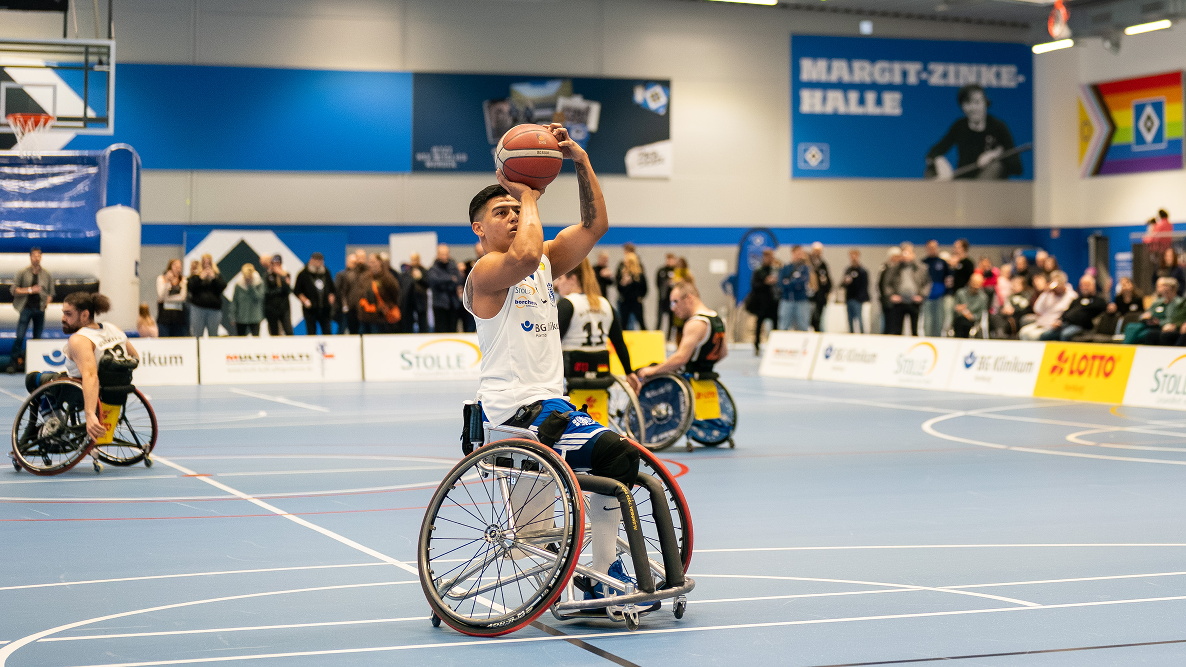 A wheelchair basketball player prepares to shoot, watched by spectators in a gymnasium, with others on the court.