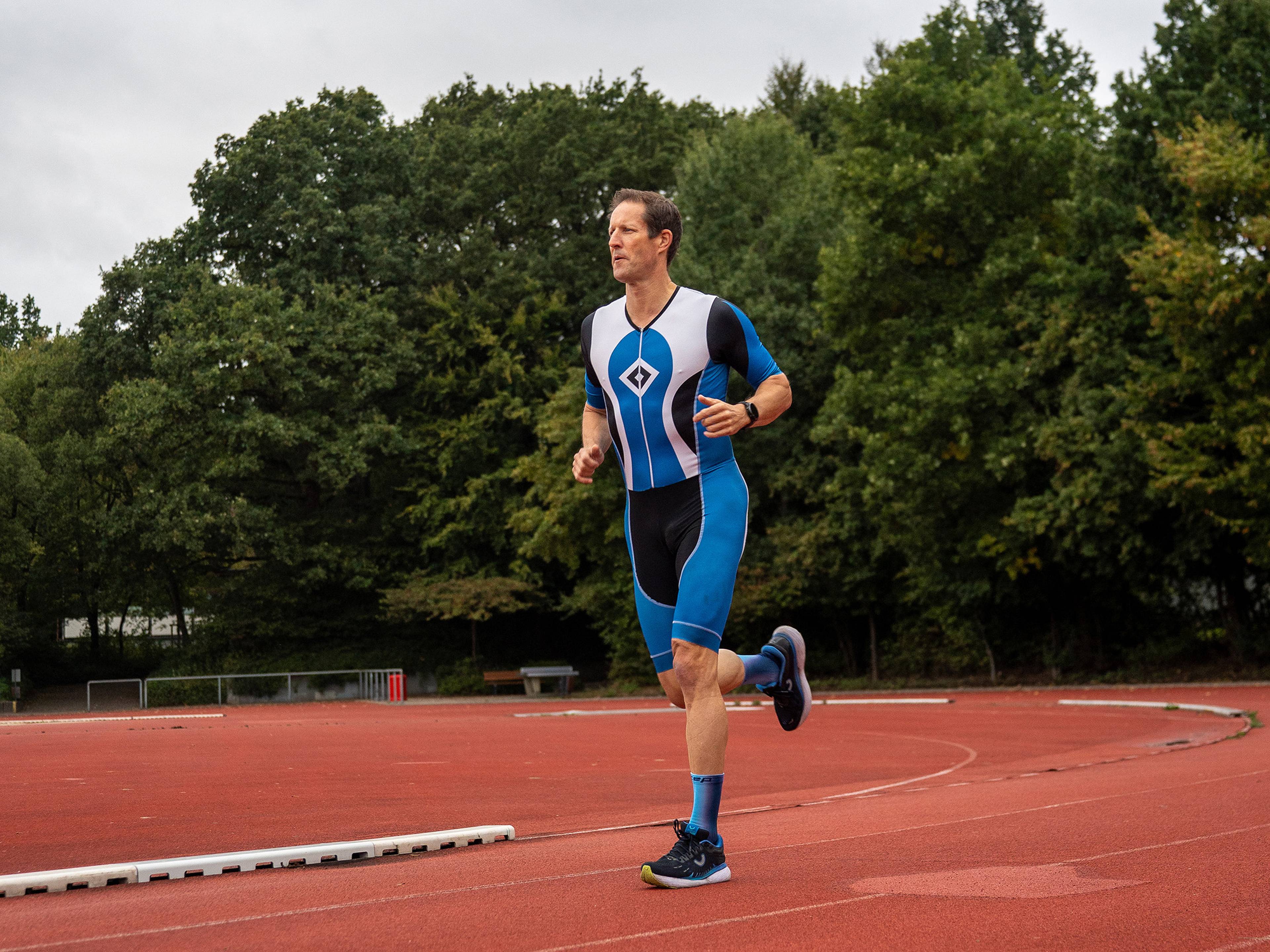 A man in a blue and white athletic outfit runs on a red track with trees in the background.