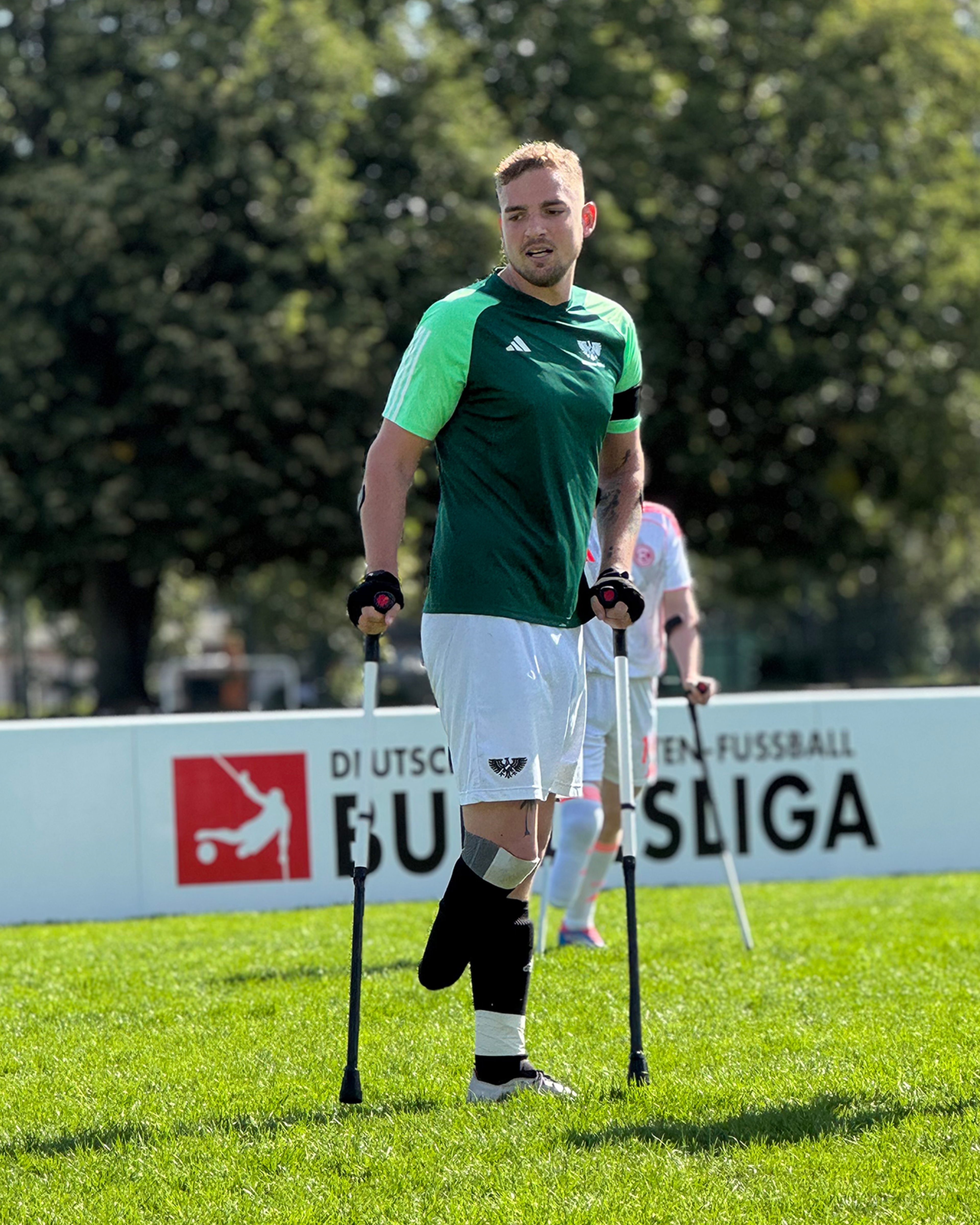 A soccer player with one leg stands on crutches on a grassy field, wearing a green jersey and white shorts, with trees in the background.