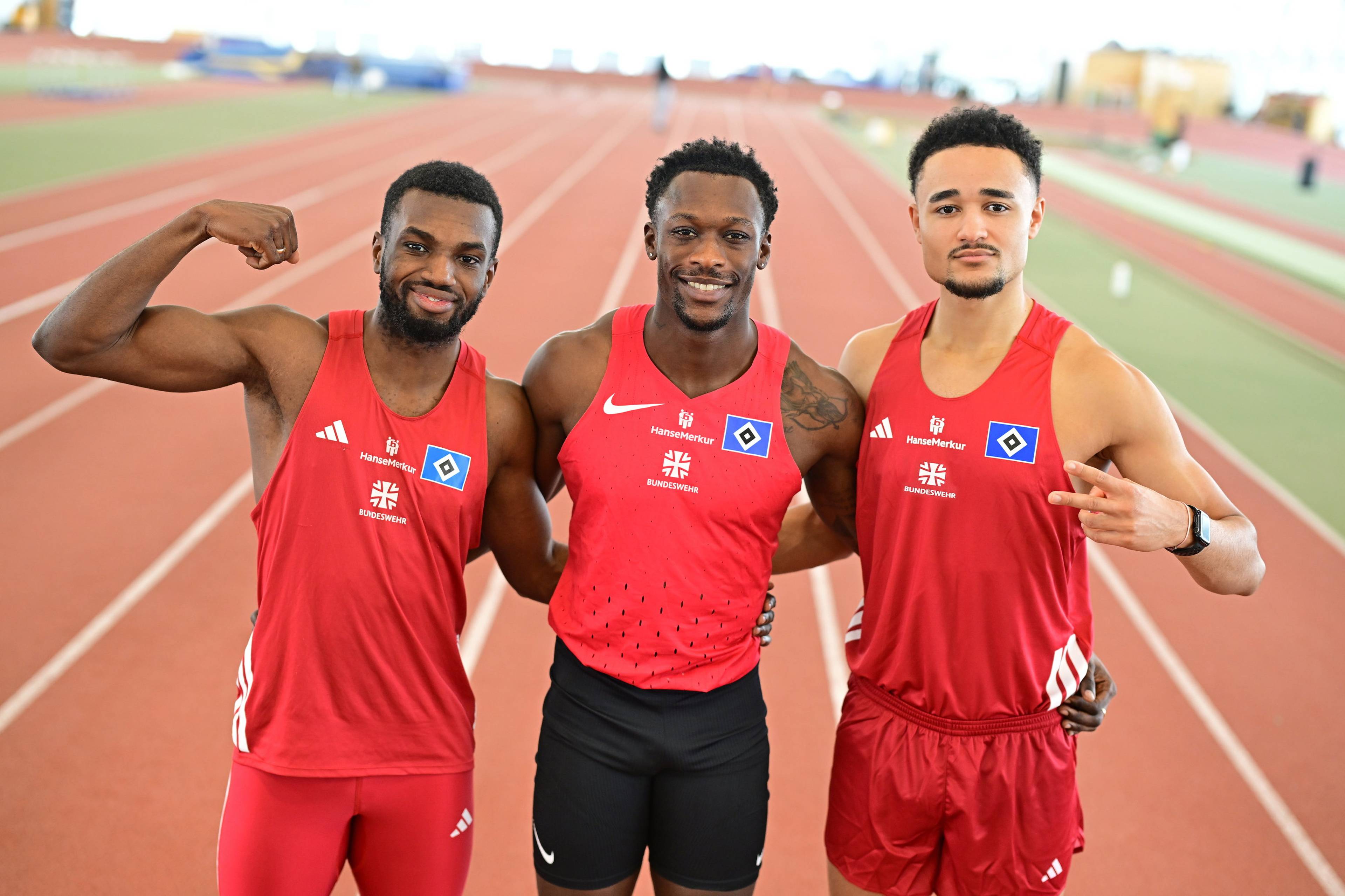 Three athletes in red track uniforms smile and pose arm-in-arm on a running track, with one flexing his arm muscle.