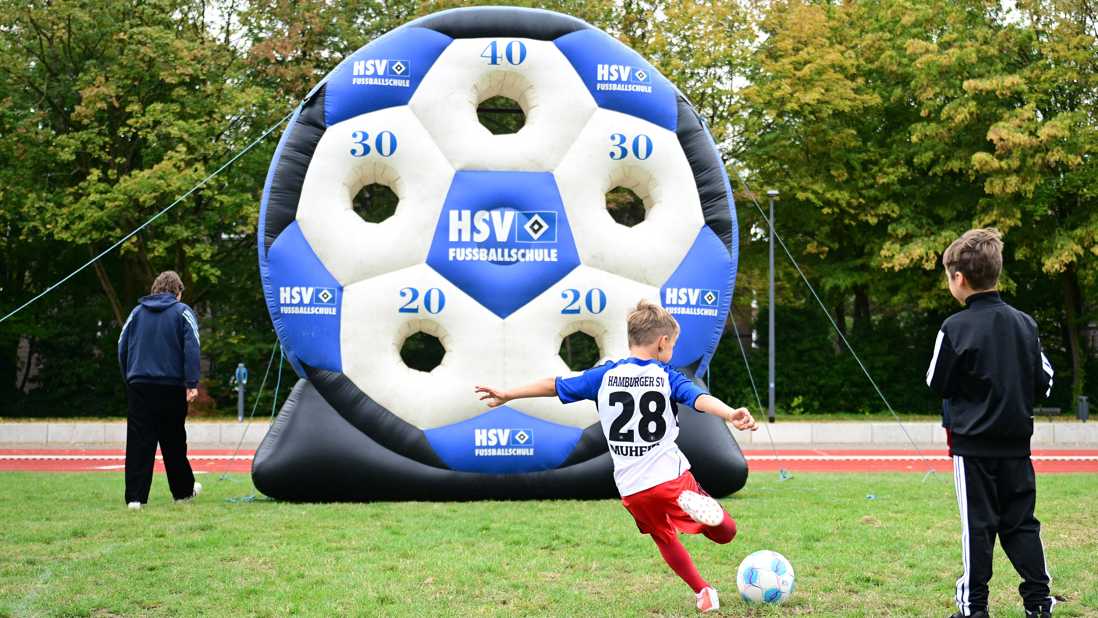 A boy in a soccer jersey kicks a ball towards an inflatable target with numbered holes, while another child and an adult watch on a grassy field.