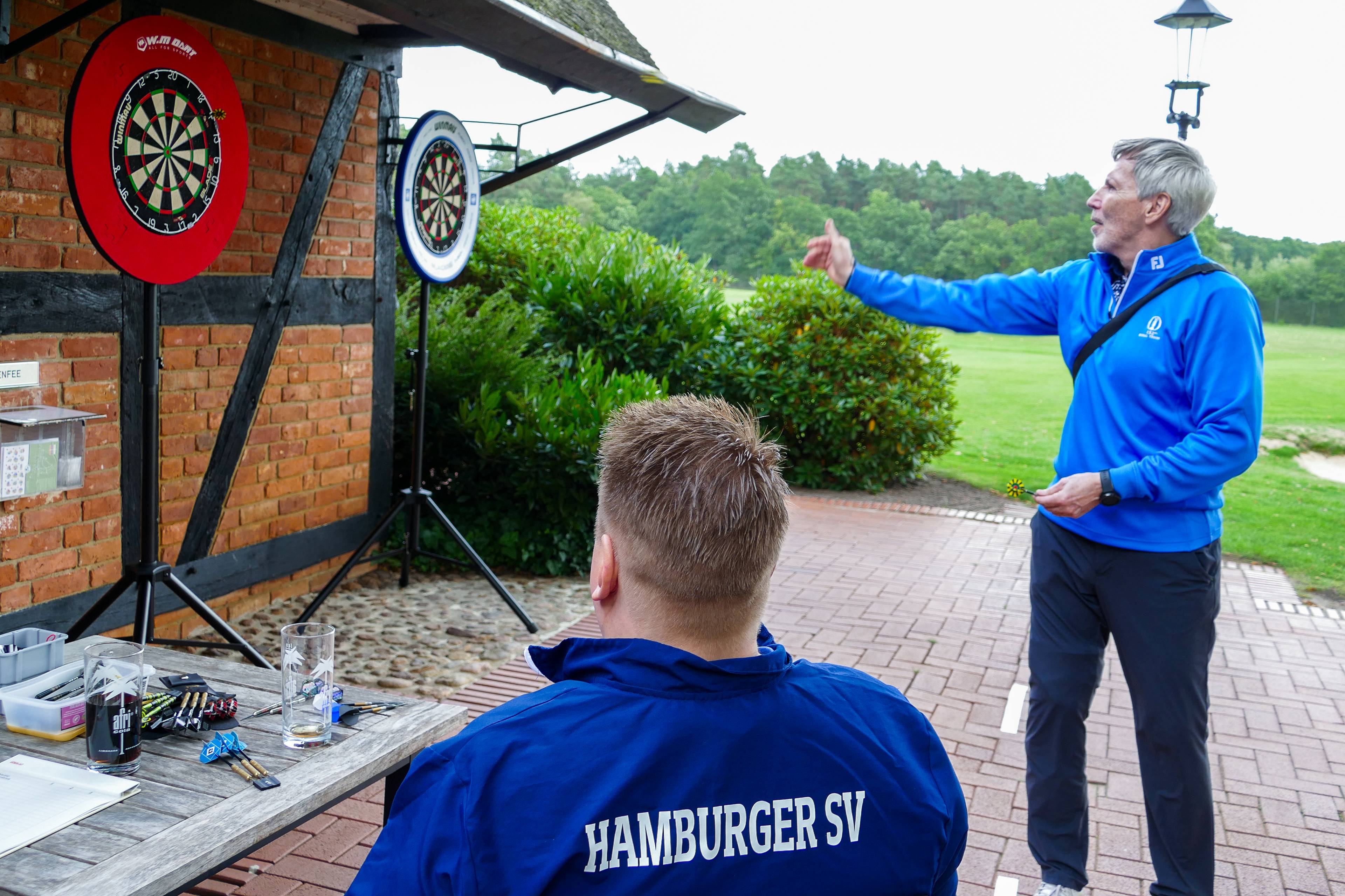 Two men playing darts outdoors, one seated with "Hamburger SV" on his jacket, the other throwing a dart towards two boards mounted on a wall.