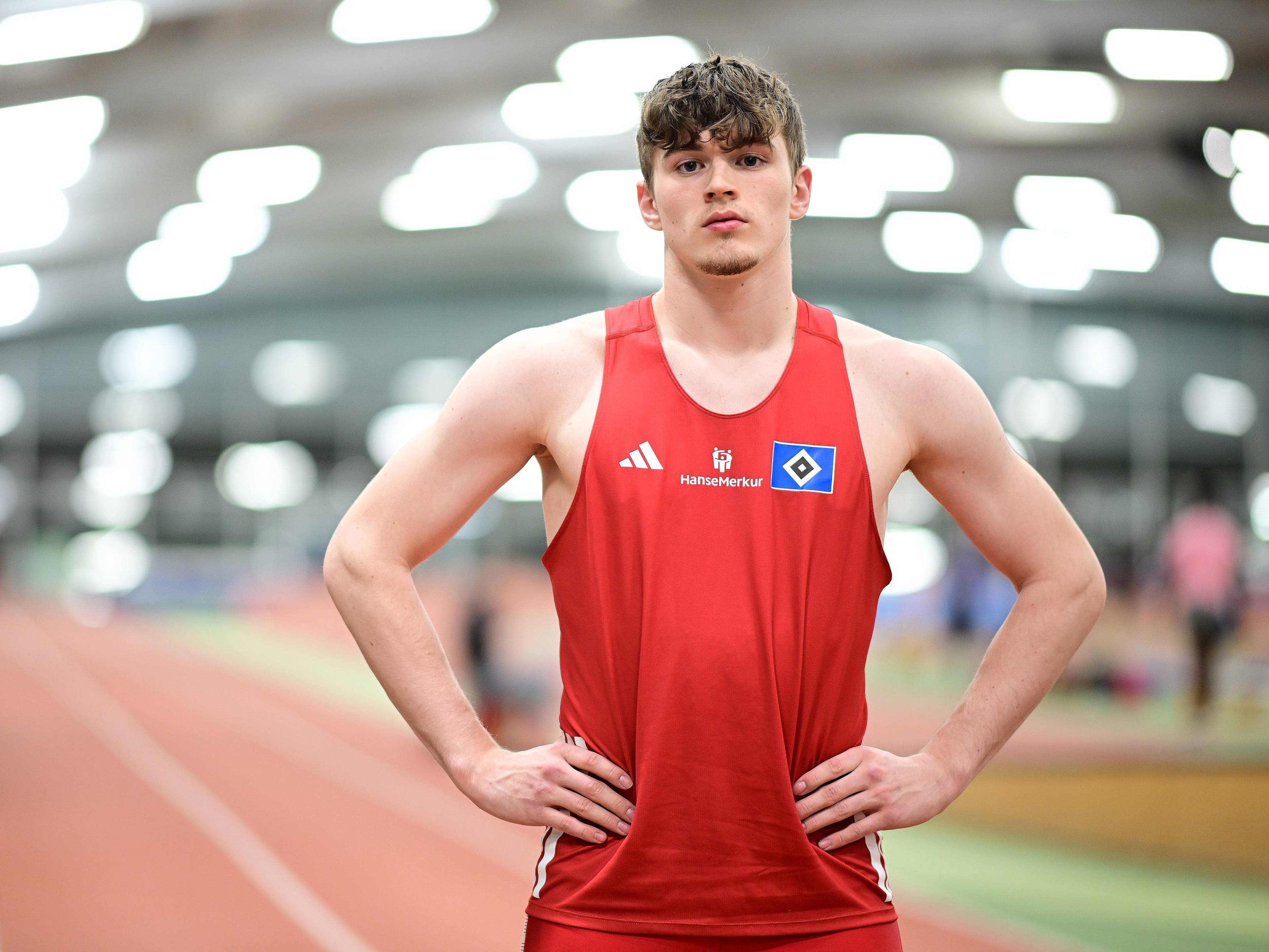 Athlete in a red sleeveless jersey stands confidently on an indoor track with hands on hips, blurred lights in the background.