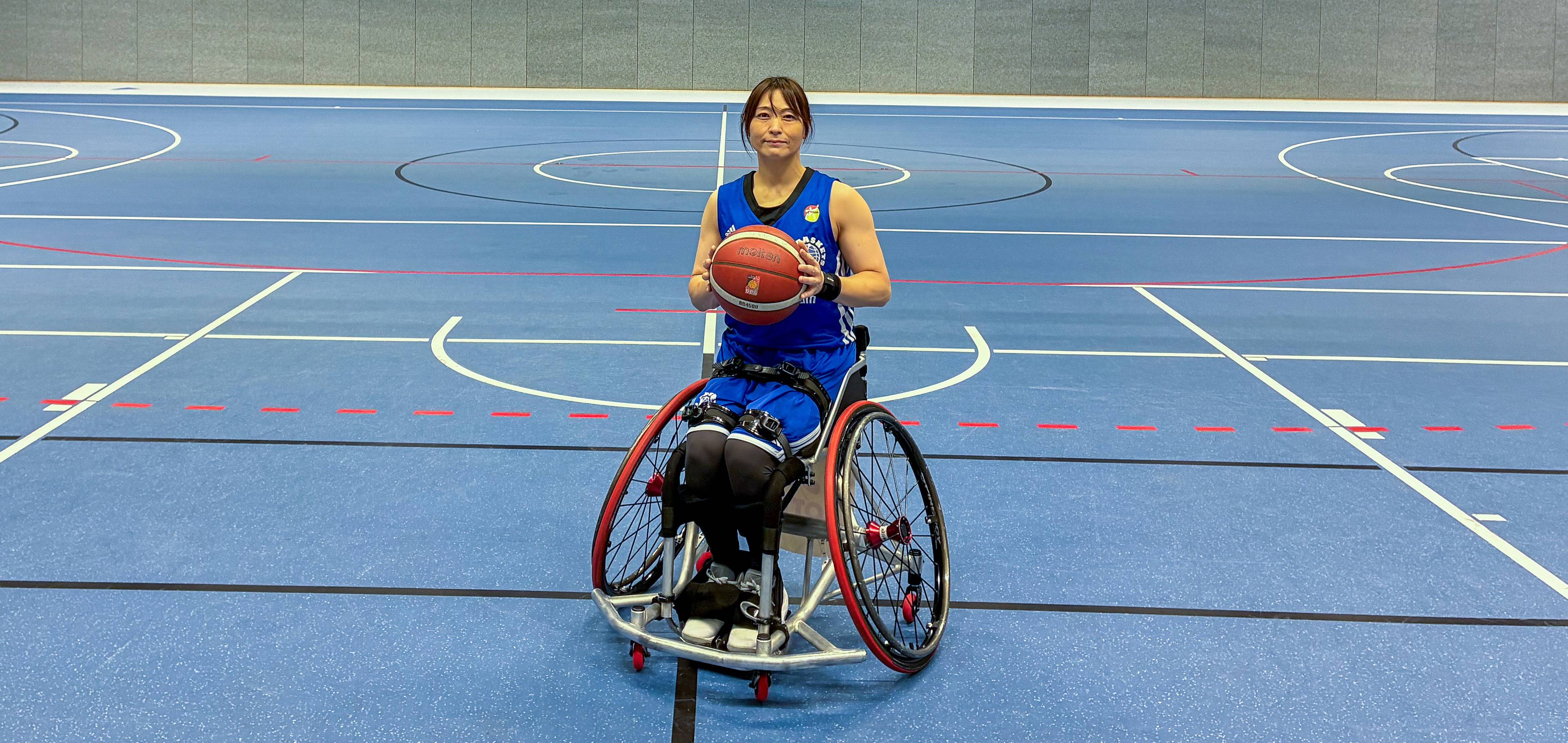 A woman in a blue jersey holds a basketball while sitting in a wheelchair on an indoor basketball court.