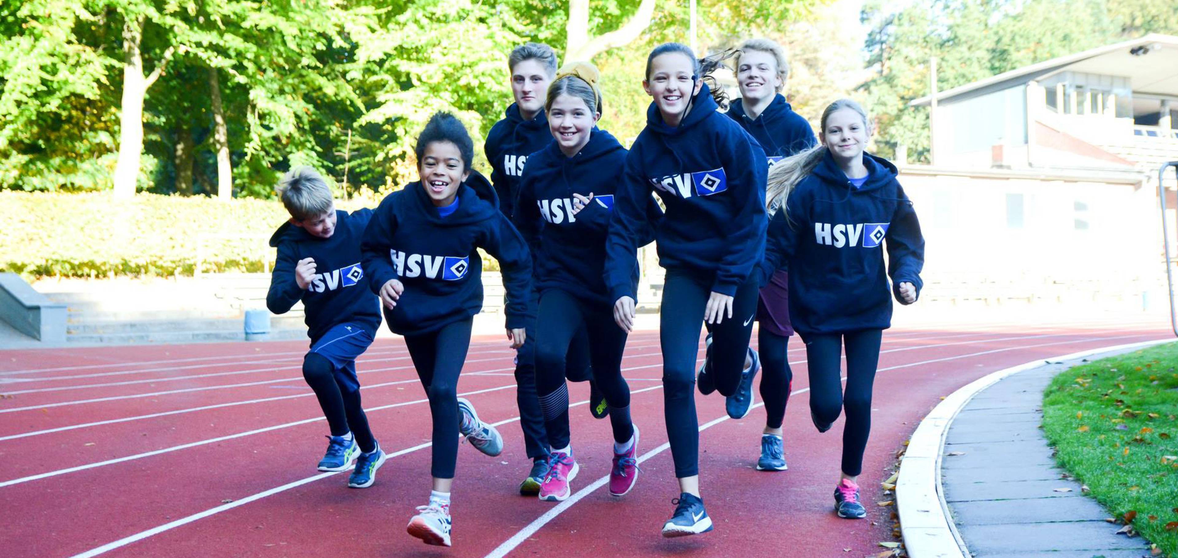 A group of smiling children in matching "HSV" hoodies run together on a track, surrounded by greenery and a few buildings in the background.