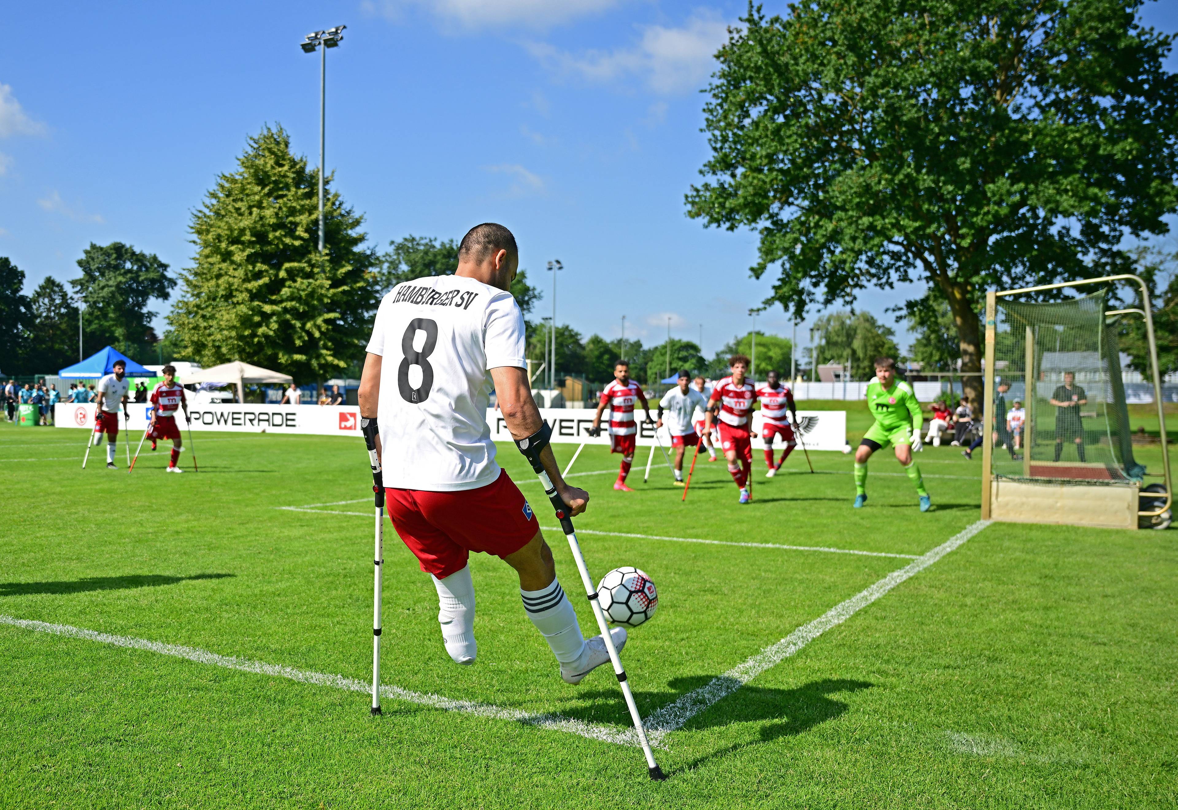 A player on crutches wearing jersey number 8 executes a corner kick during an amputee soccer match, with other players and a goalie in the background.