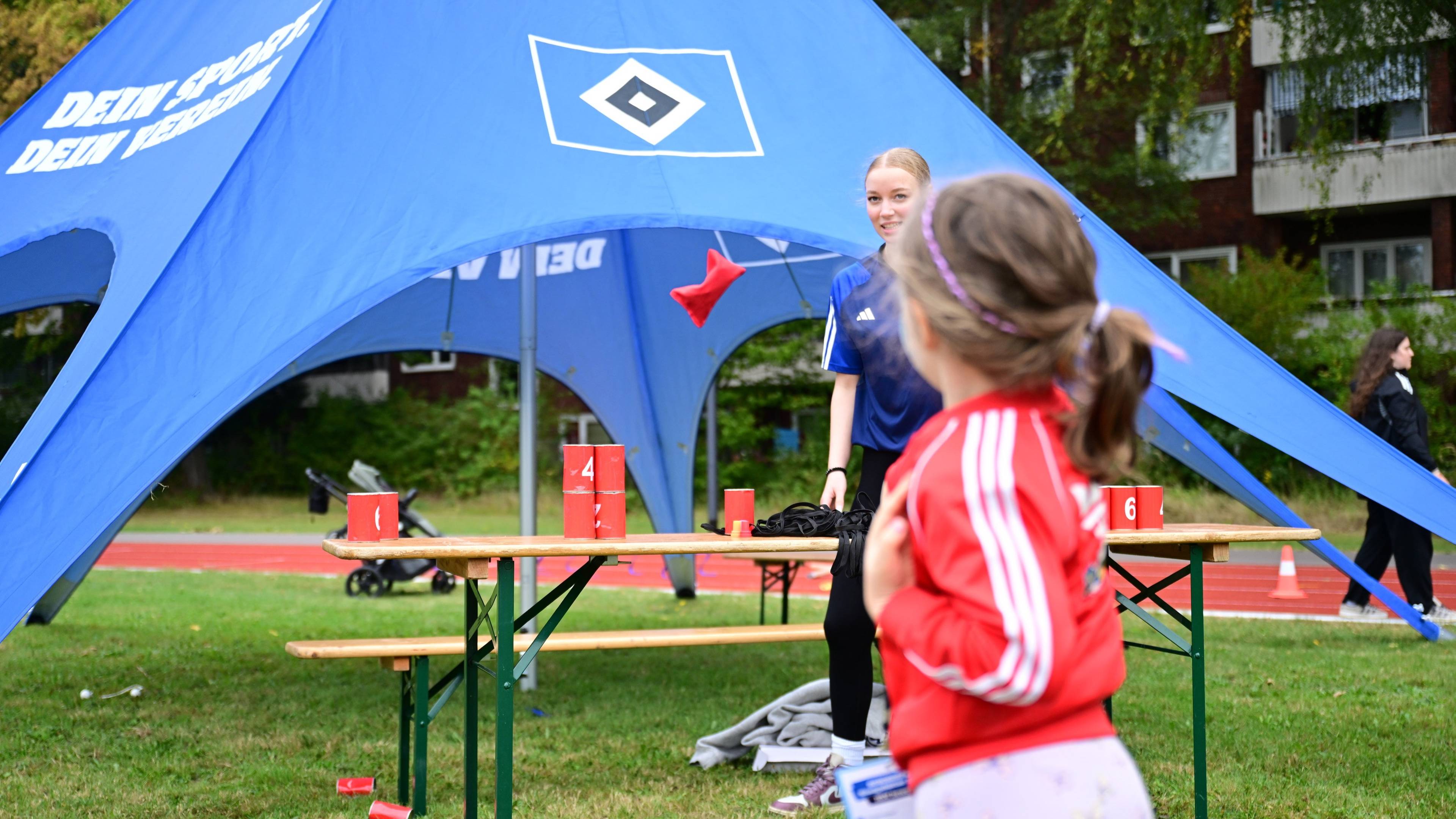 Child in red jacket throws a red beanbag towards cans on a table under a blue tent at an outdoor event.
