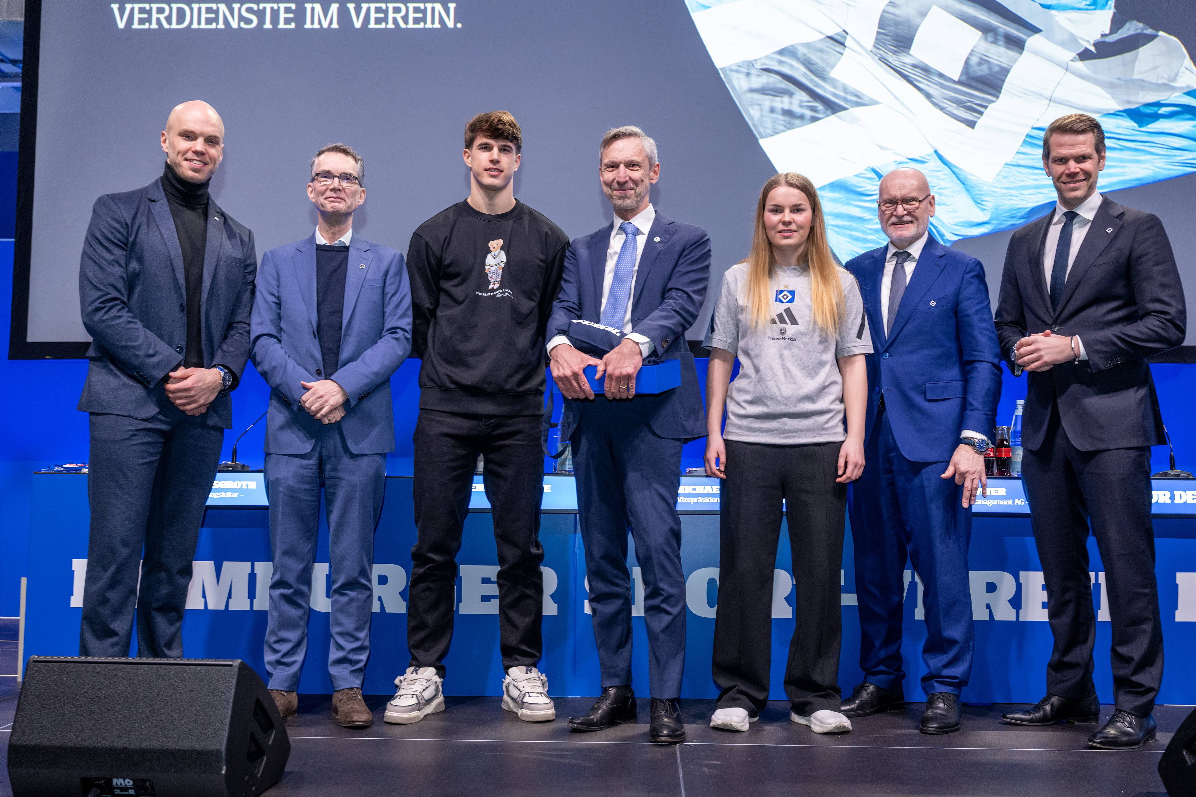 Seven people stand on stage in formal attire, with a backdrop displaying a flag and German words.
