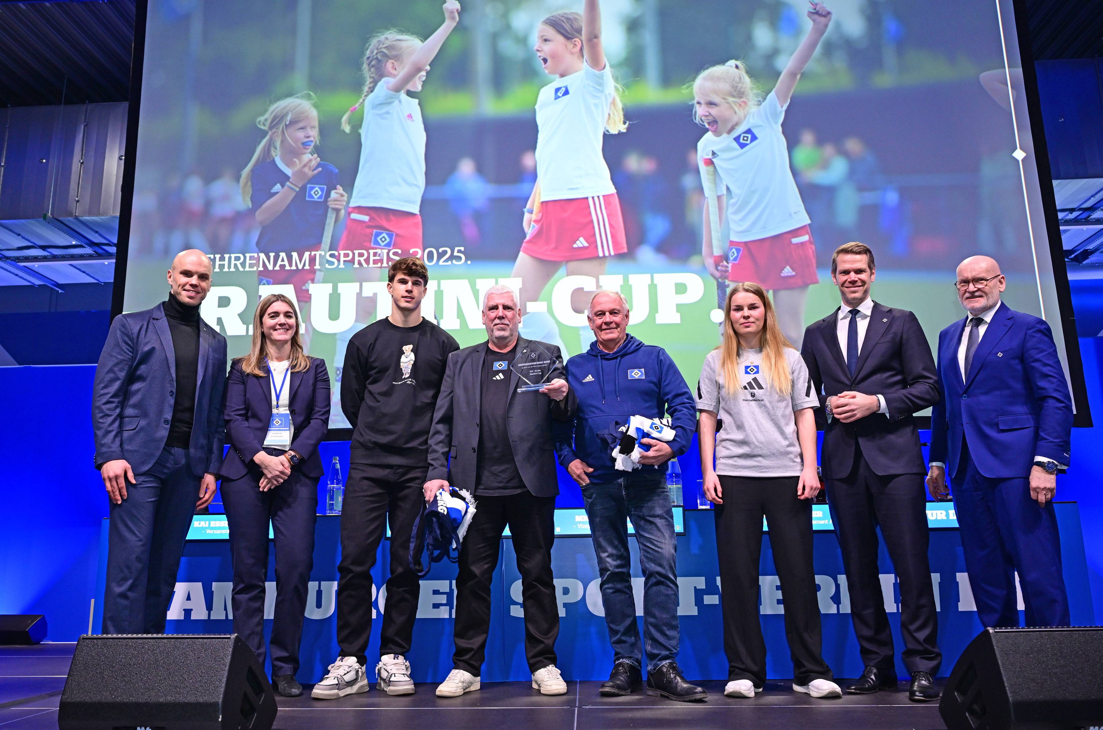 A group of eight people, smiling on stage in front of a large screen showing children playing, during an event with a 2025 sports award theme.