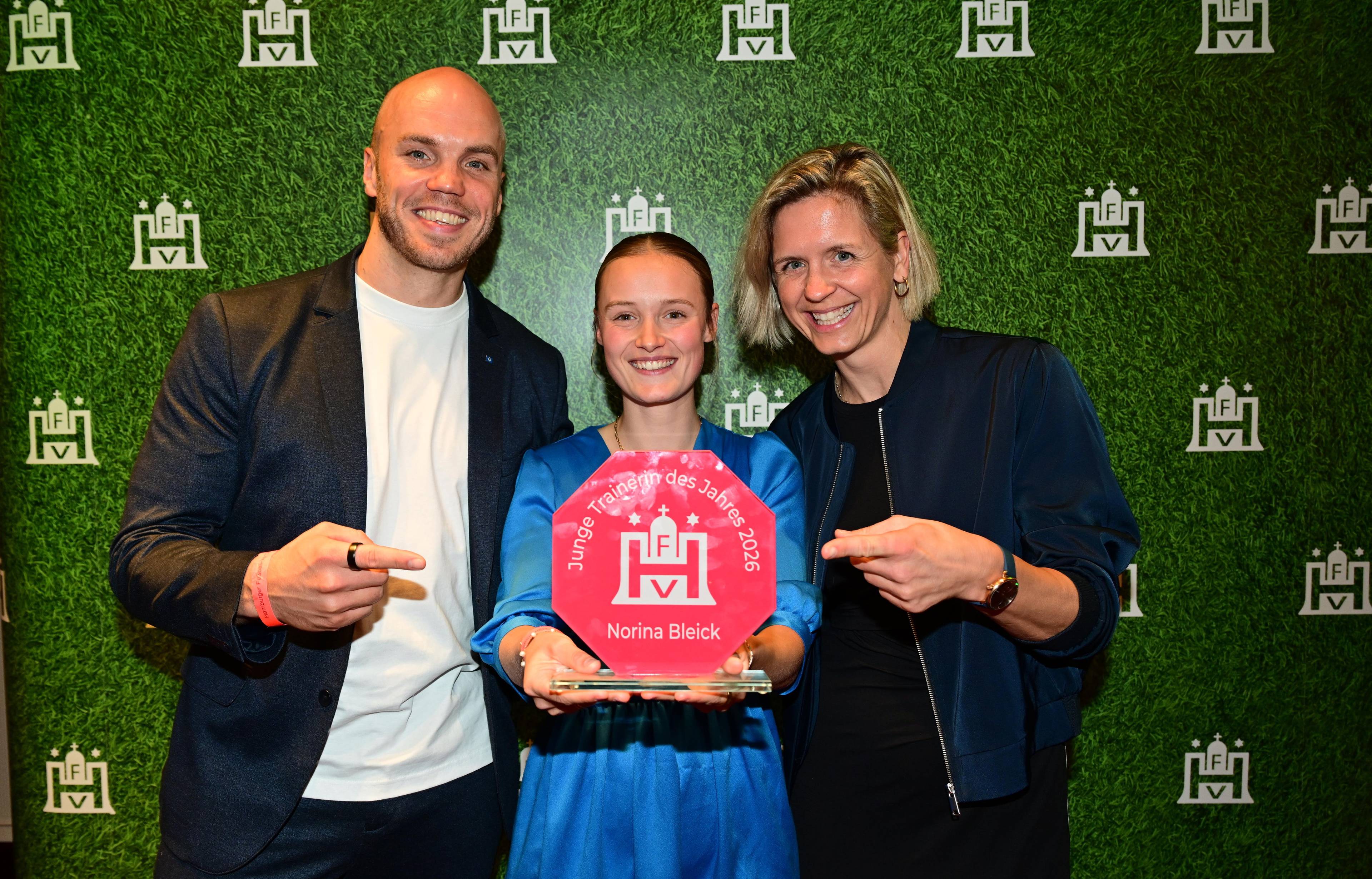 Three people smiling, holding an award with the text "Junge Trainerin des Jahres 2026" against a green backdrop with white logos.