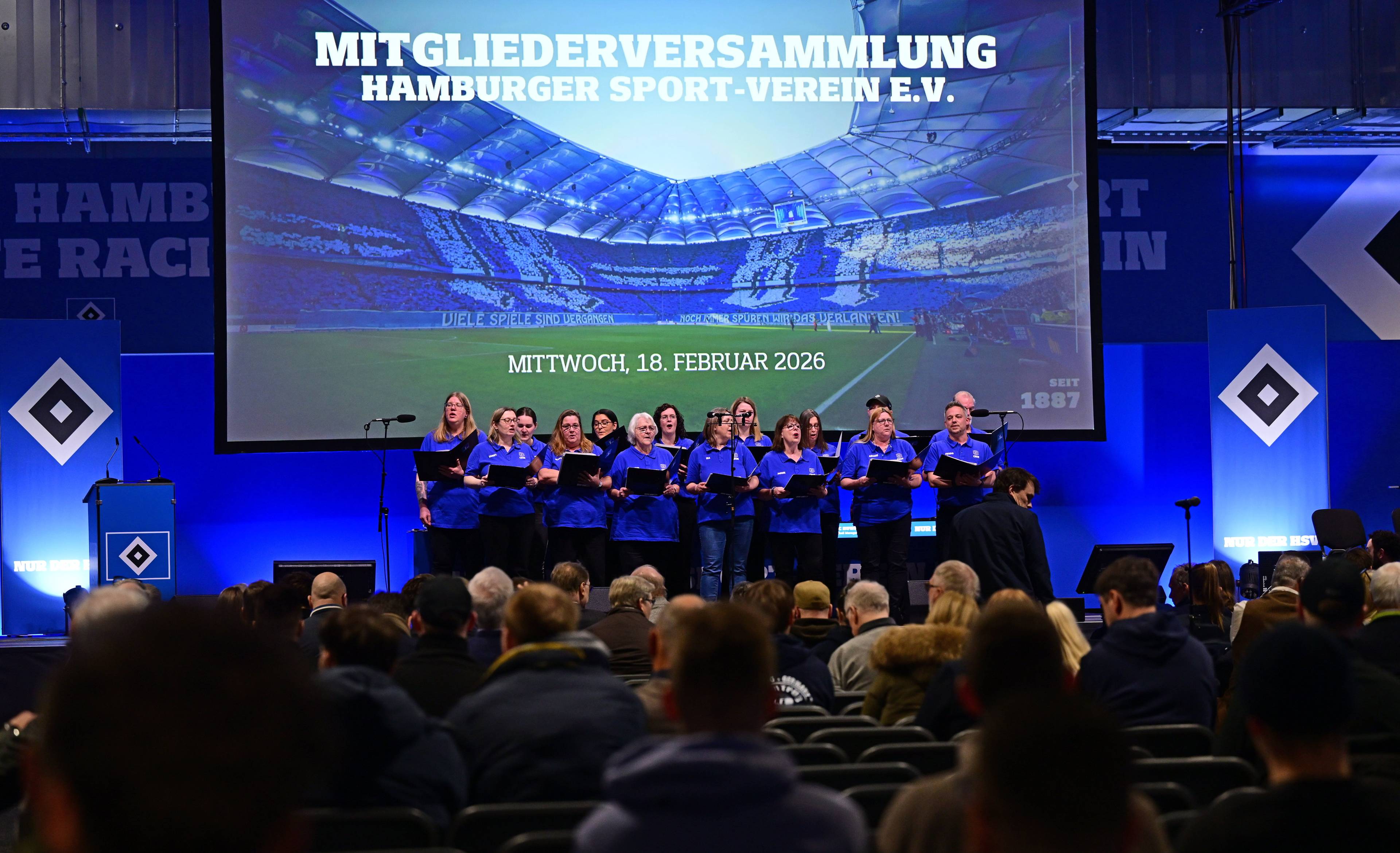 A group of people stands on stage singing, with a large screen in the background displaying event details for a sports club meeting.