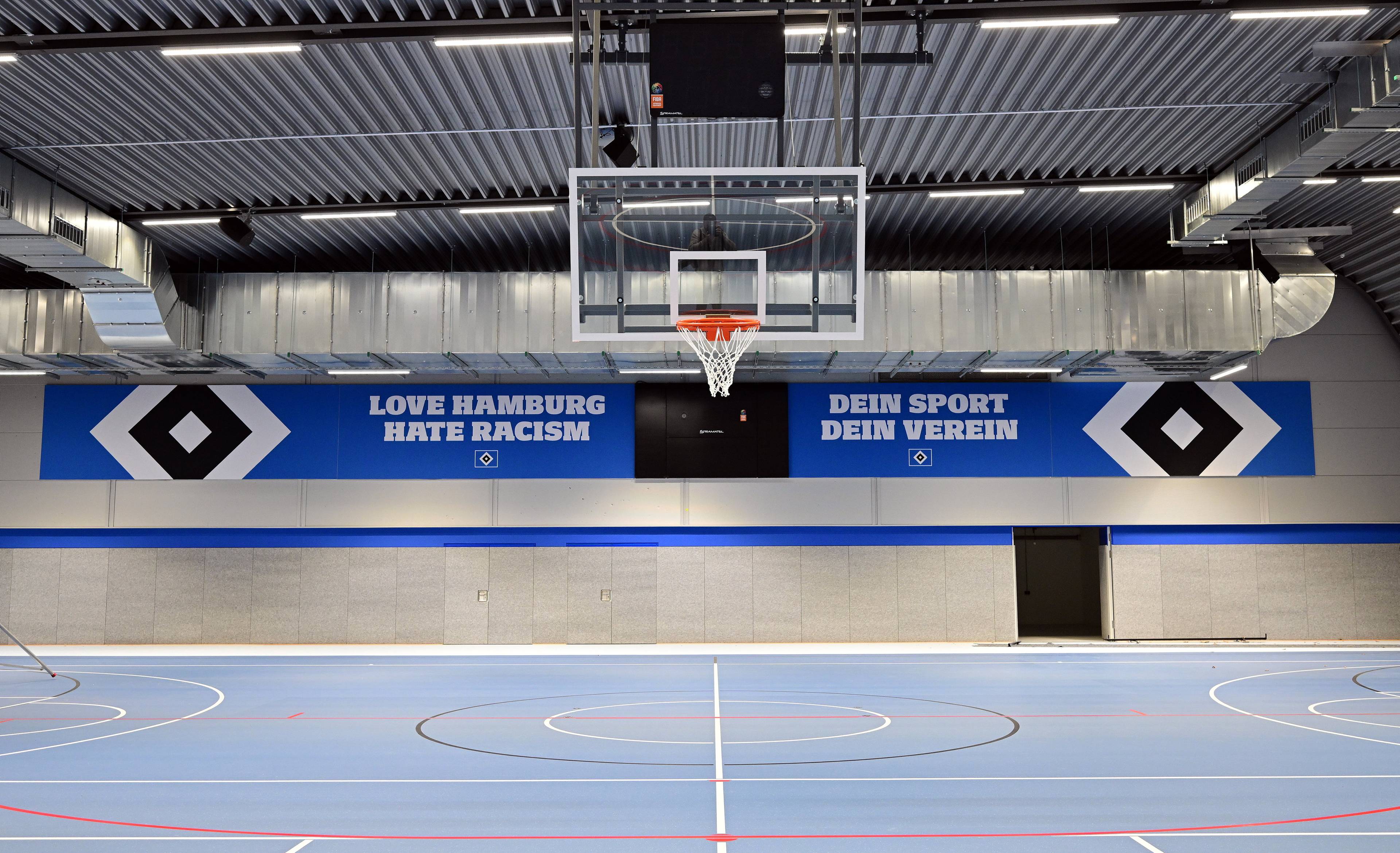 Indoor sports hall with a basketball hoop, blue and gray walls, and banners reading "Love Hamburg, Hate Racism" and "Dein Sport, Dein Verein."