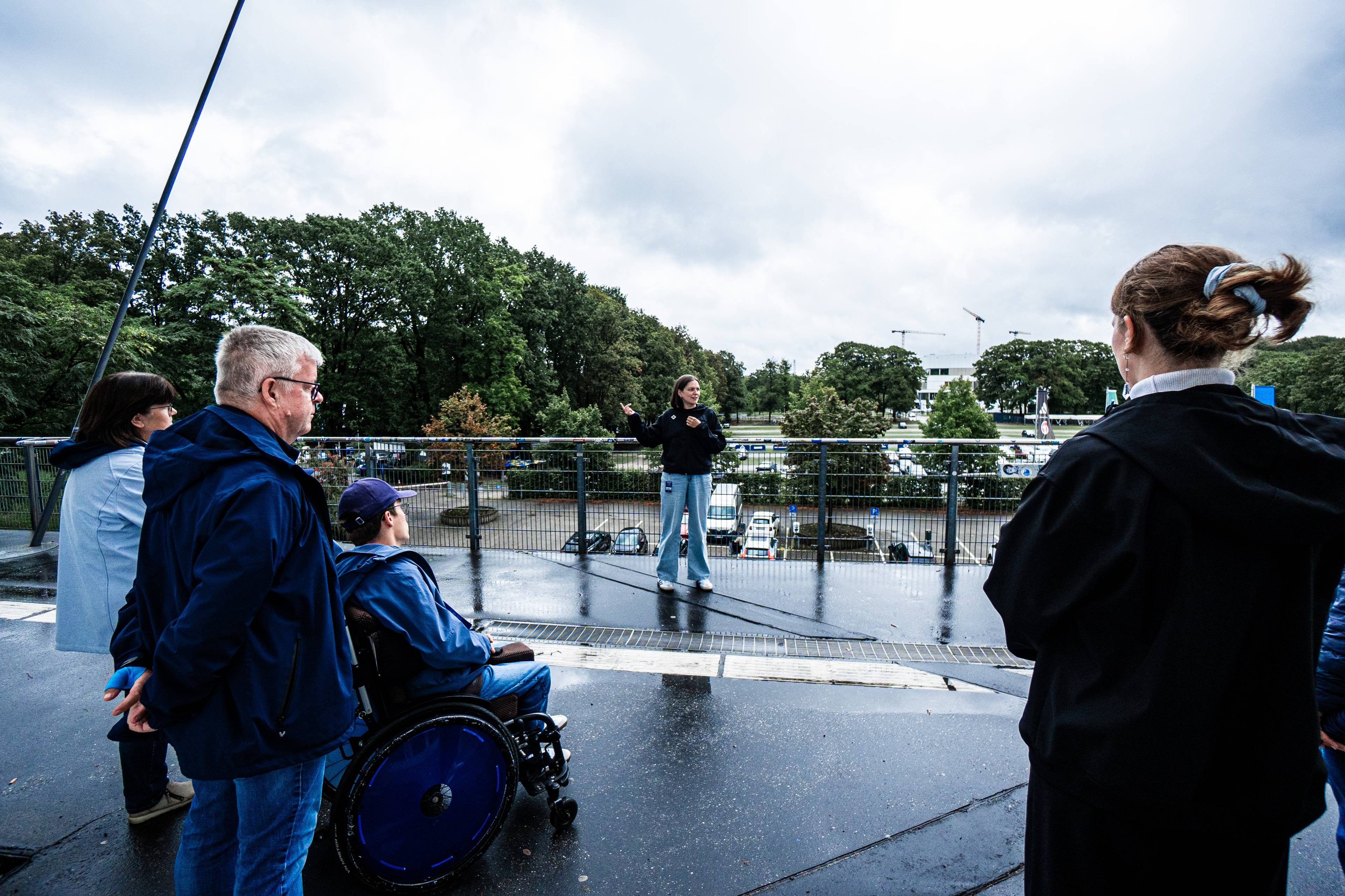Eine Gruppe von Menschen, darunter eine Person im Rollstuhl, lauscht an einem regnerischen Tag einer Rednerin, im Hintergrund sind Bäume und geparkte Autos zu sehen.