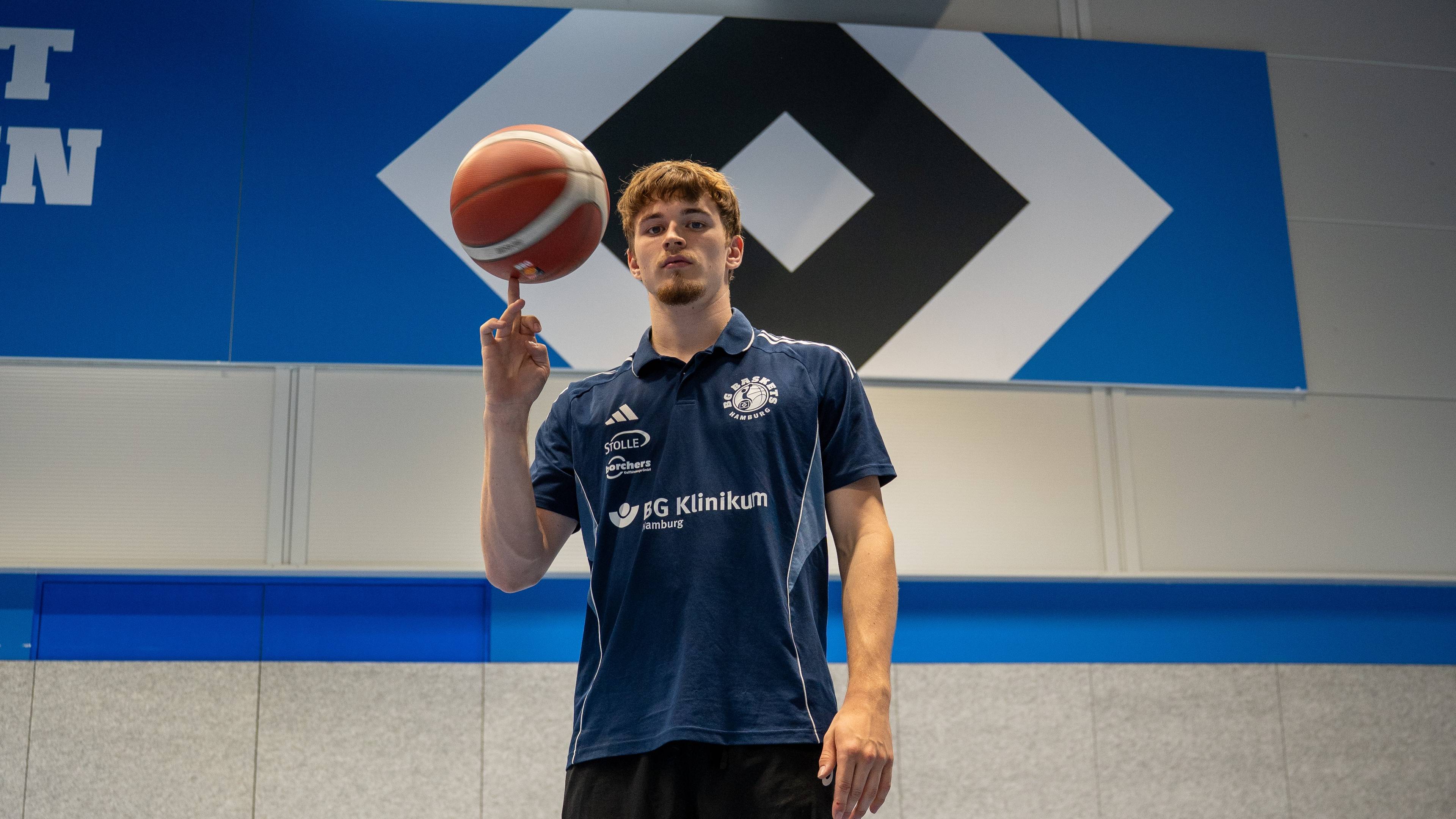 A man in a blue shirt with logos spins a basketball on his finger indoors with a large geometric poster background.