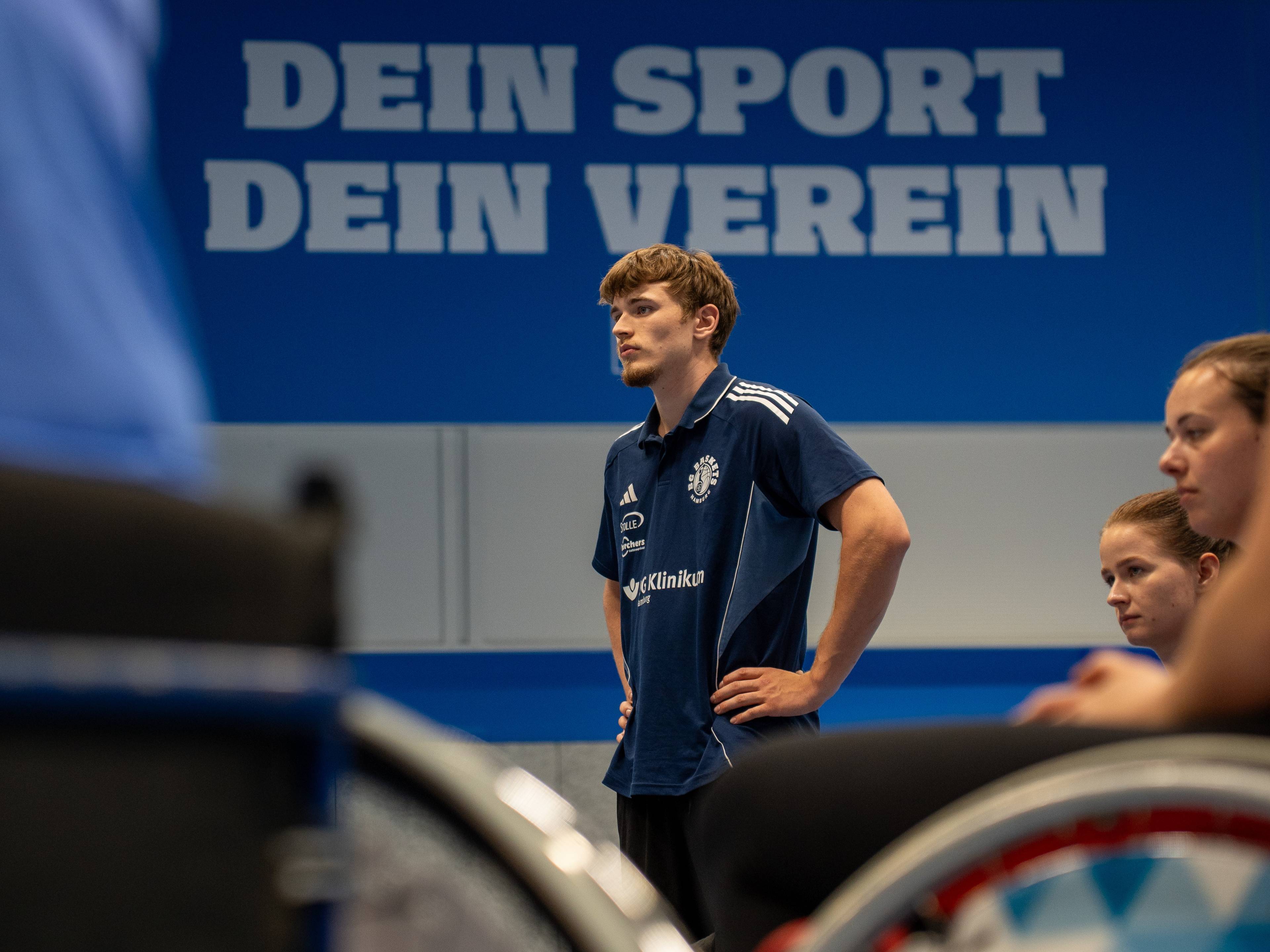 A man in a sports jersey stands with hands on hips, several people seated nearby, and "Dein Sport Dein Verein" in large letters on the wall.