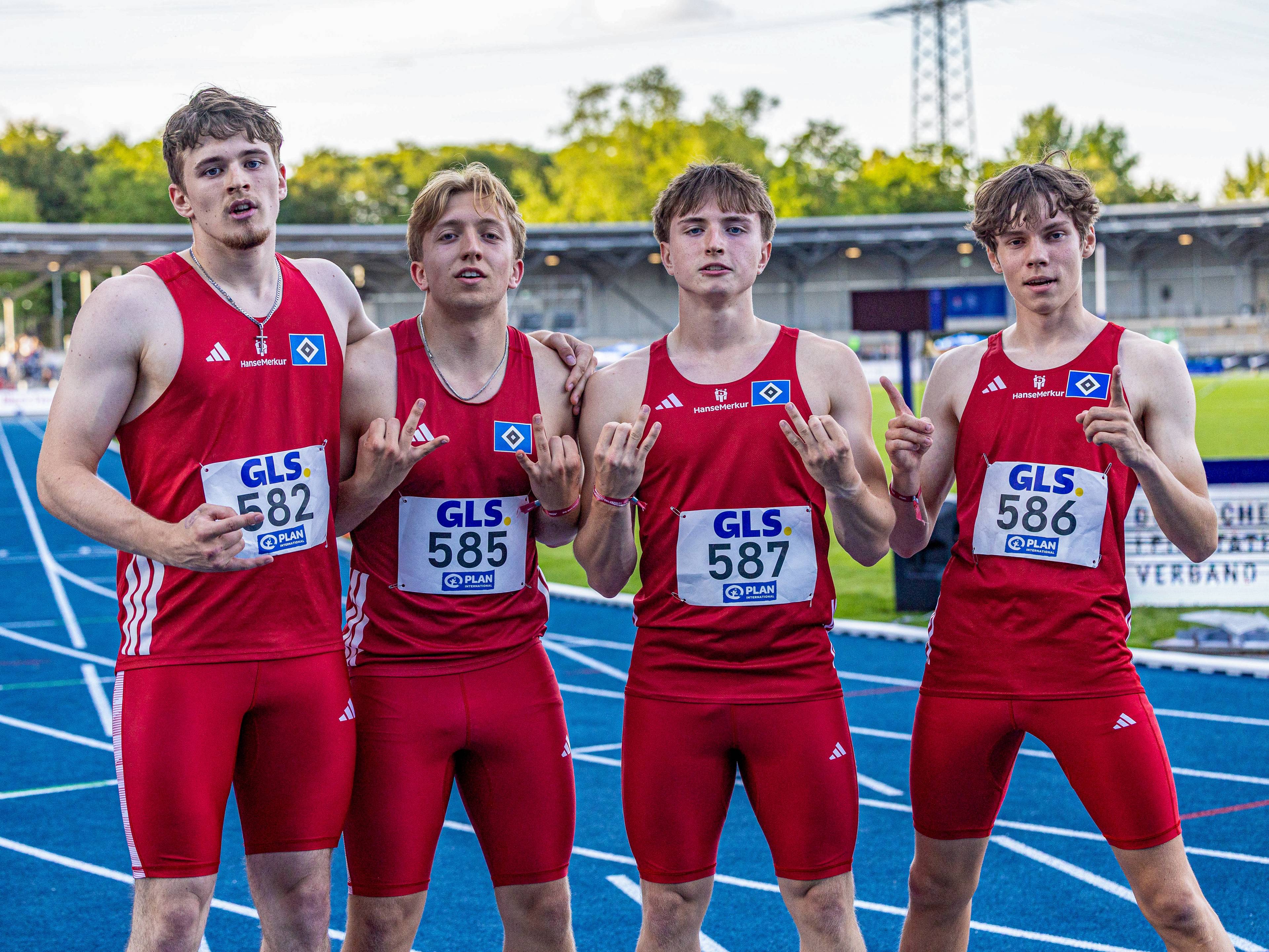 Four male athletes in red uniforms with race numbers, posing on a blue track at a stadium.
