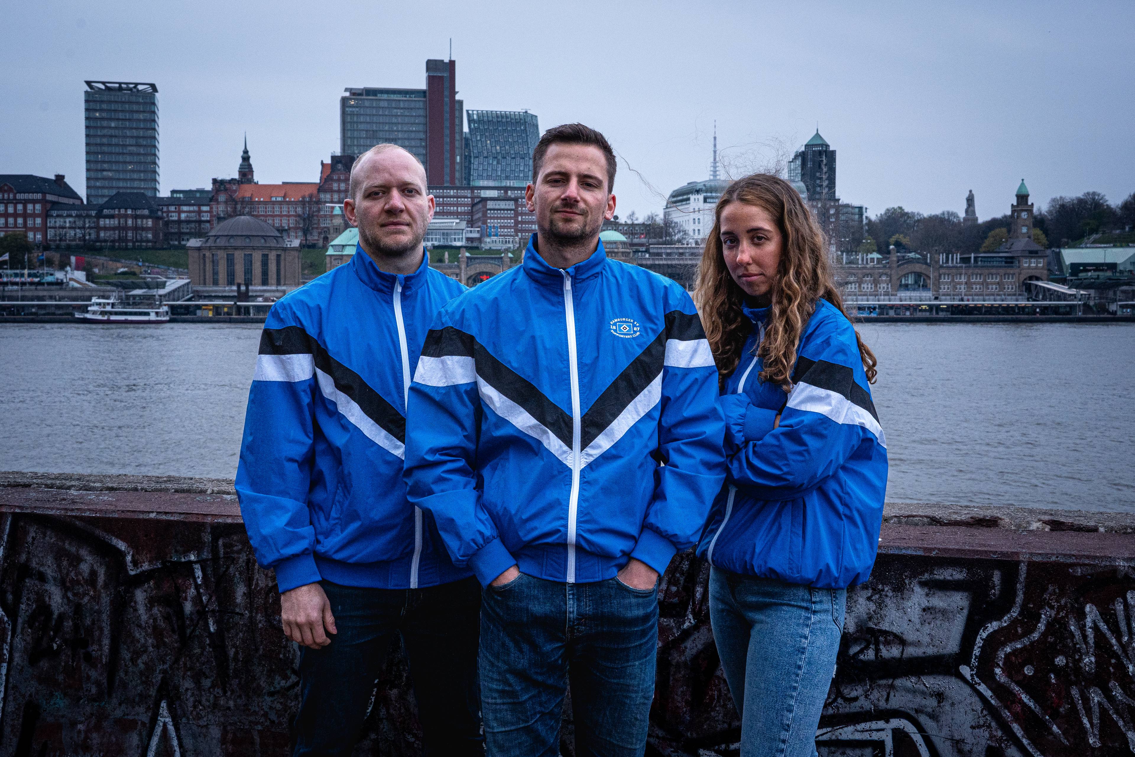 Three people in blue track jackets stand by a graffiti-covered railing with a city skyline and waterfront in the background.