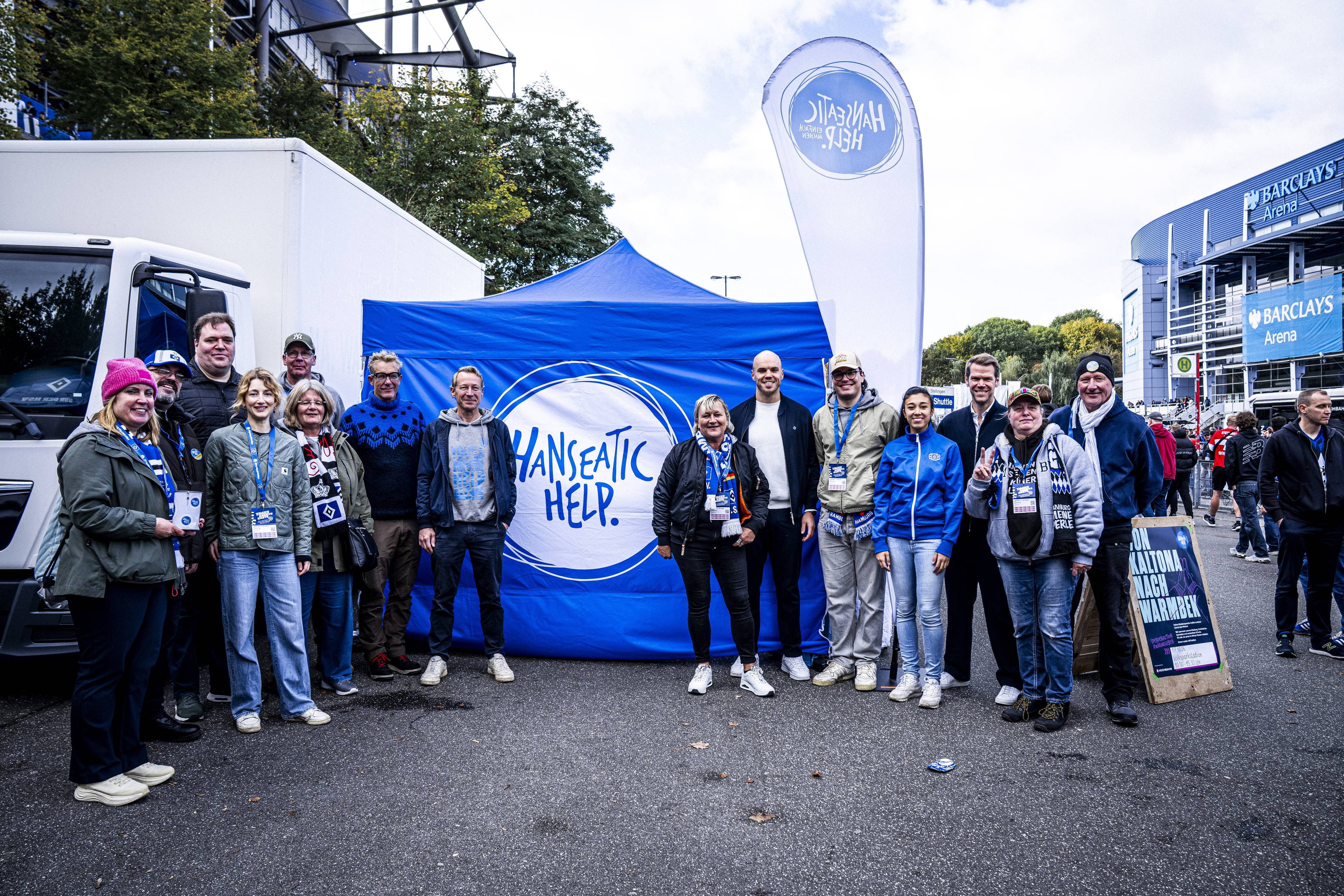 Eine Gruppe von Menschen vor einem Pavillon mit der Aufschrift "Hanseatic Help". Im Hintergrund sind ein weißer LKW, das Volksparkstadion und die Barclays Arena zu sehen.