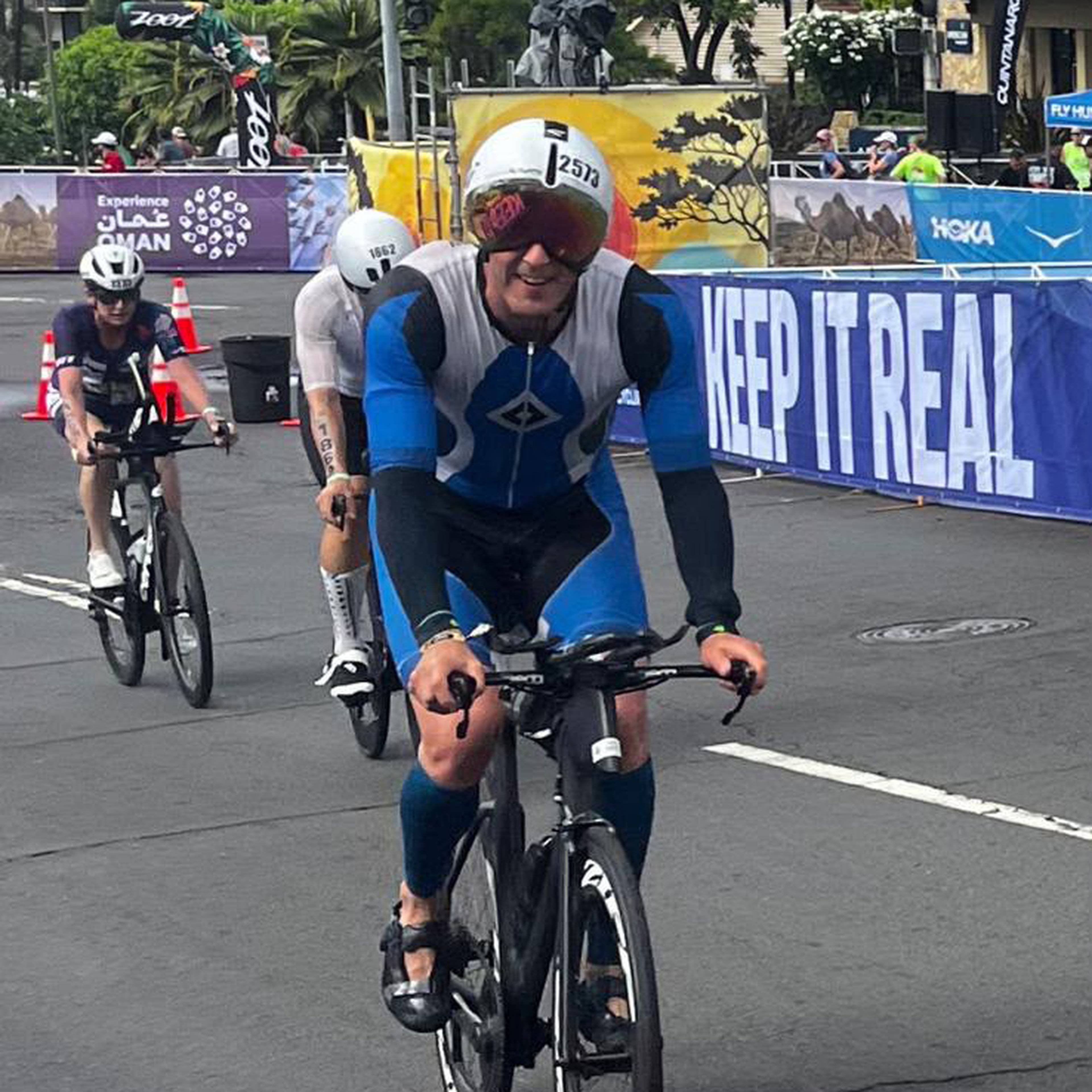 Cyclists in a race, focused and wearing helmets, approach a section with a "KEEP IT REAL" banner on the side.
