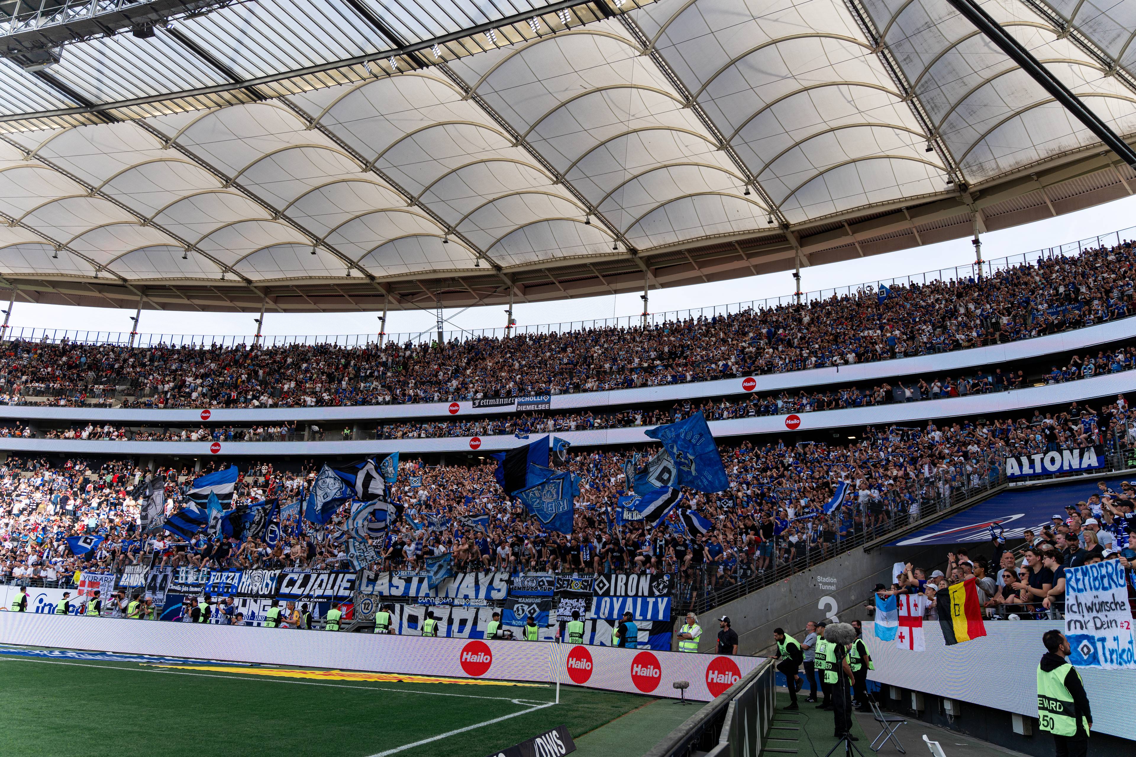 Large crowd of fans in a stadium, holding blue flags and banners, cheering enthusiastically. Security personnel stand near the field.