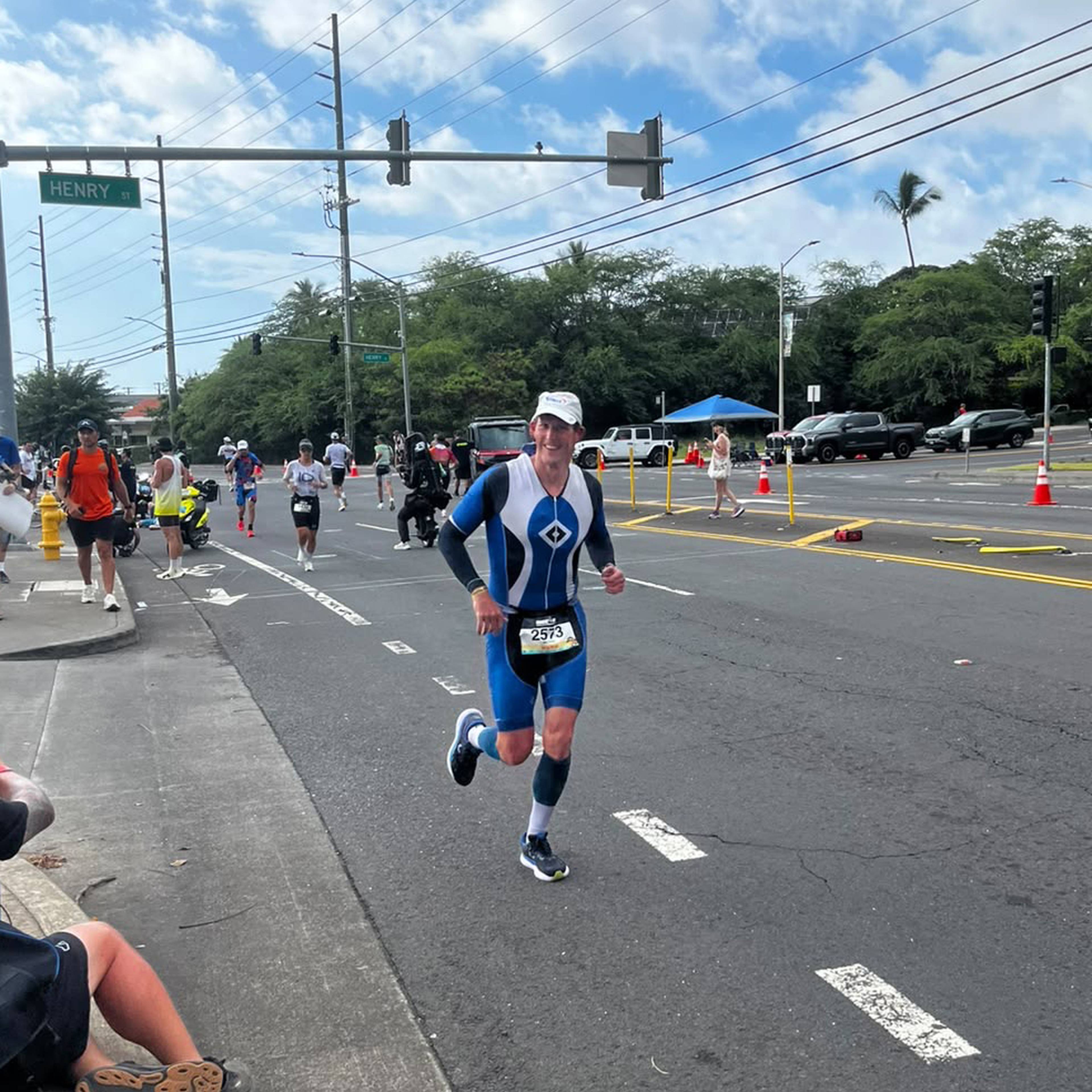 A person in a blue and white triathlon suit running on a street during a race, surrounded by spectators and other participants.