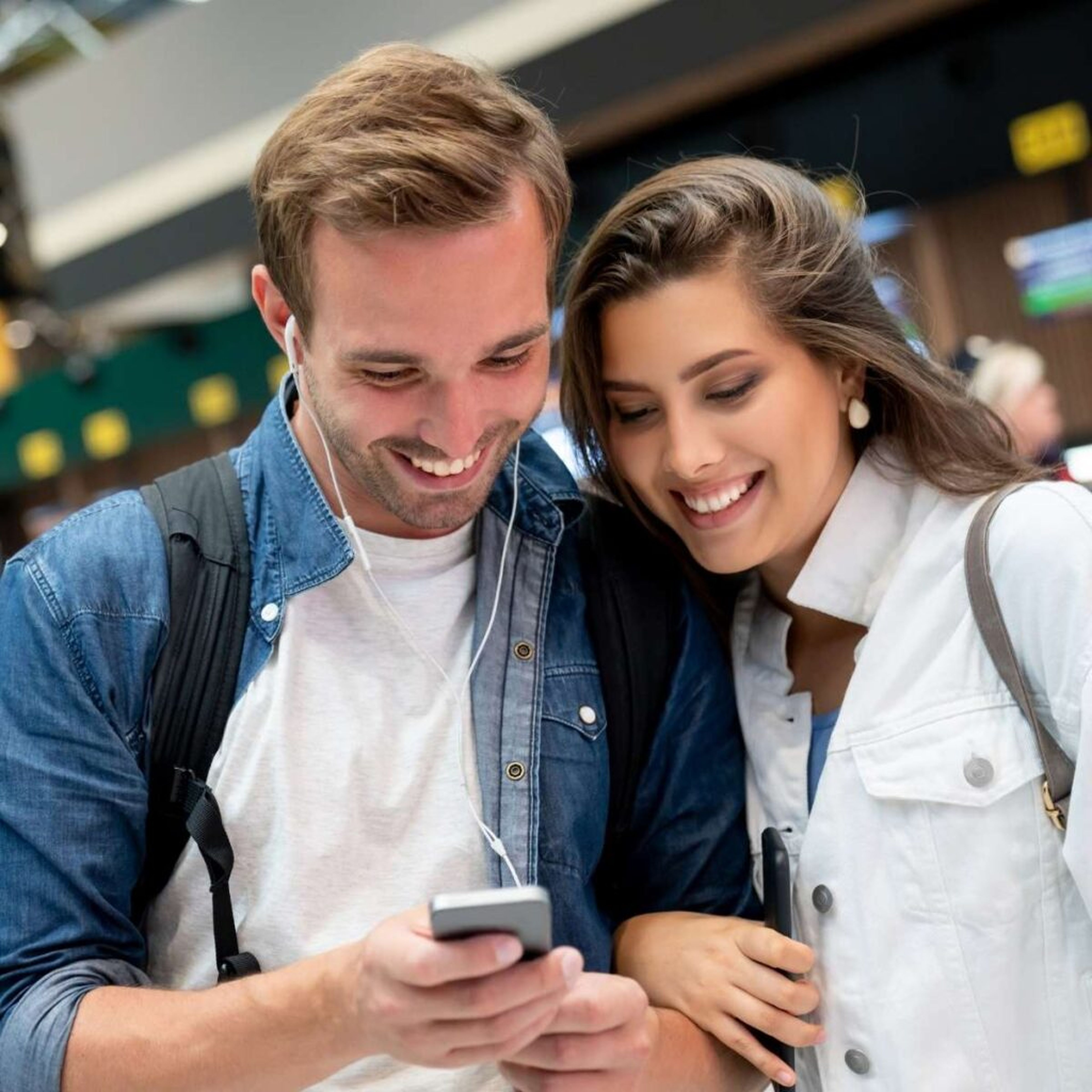Ein lächelndes Paar schaut auf ein Smartphone und trägt Kopfhörer, im Hintergrund ein Bahnhof oder Flughafen.