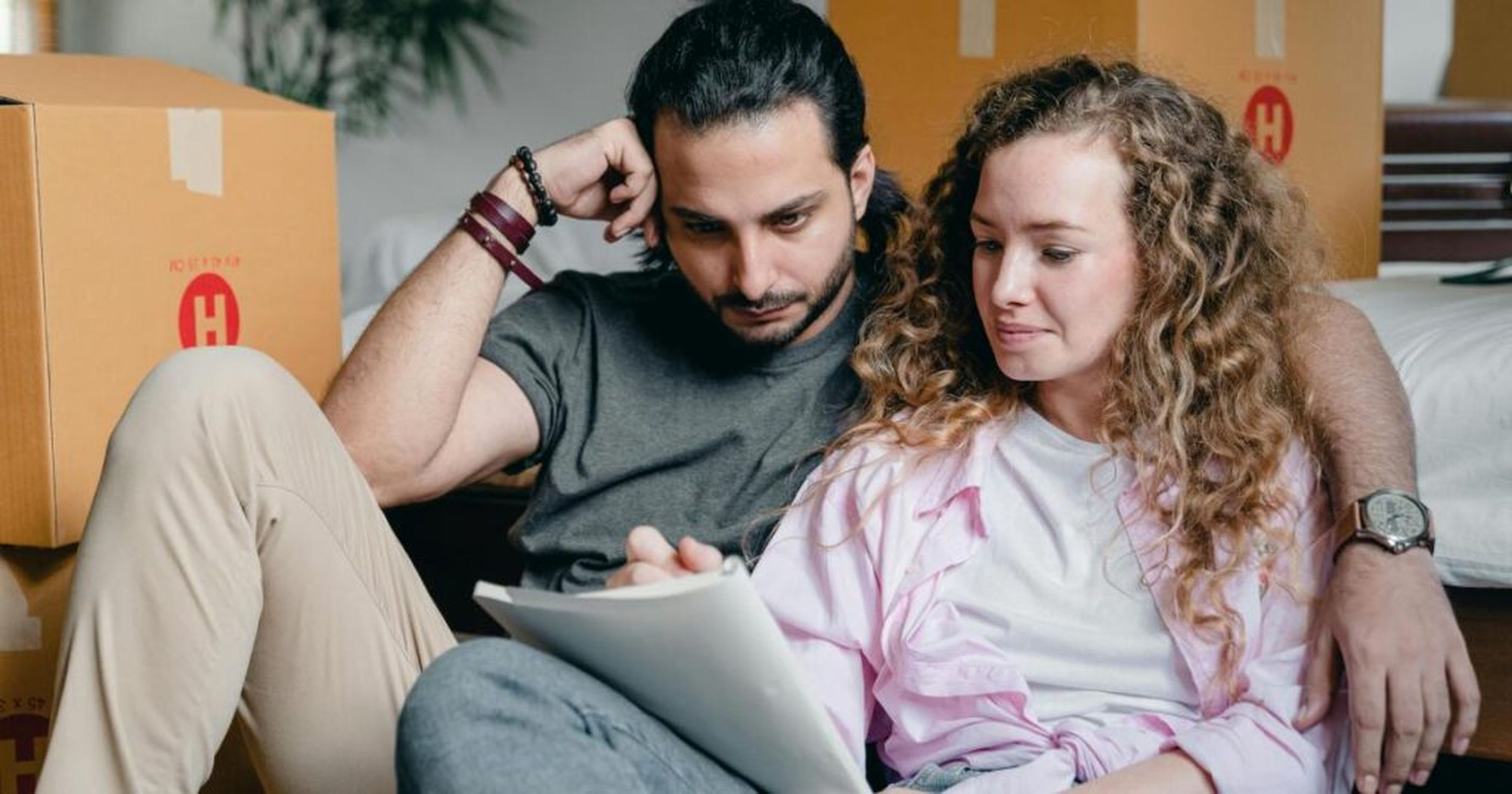 Couple sitting on floor with moving boxes, reading a document together.