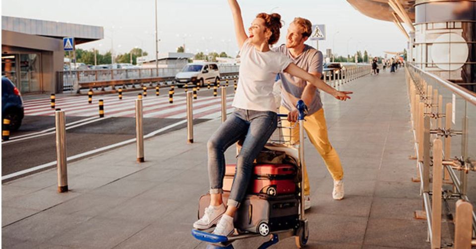 Personen am Flughafen mit Koffern, eine Frau sitzt auf einem Gepäckwagen, strahlend und die Arme ausgebreitet.