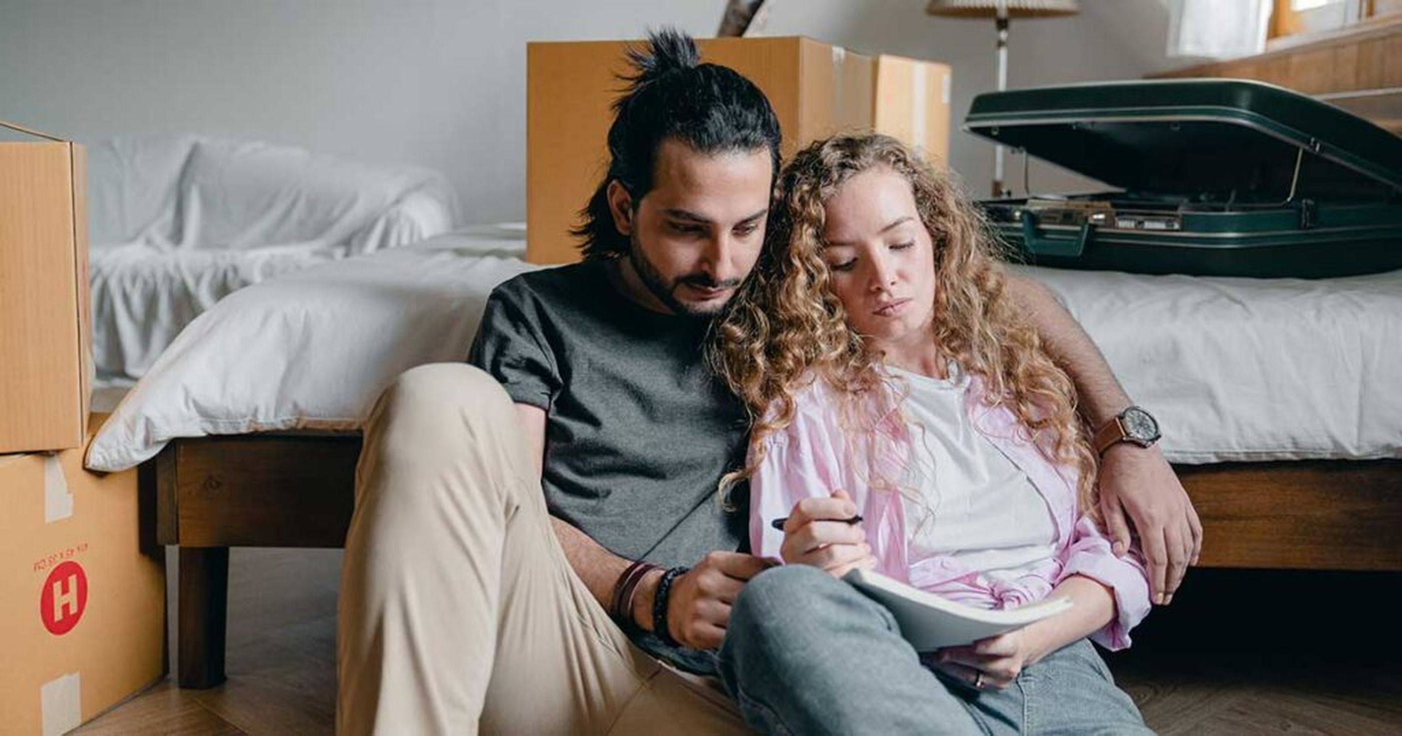 Couple assis sur le sol, entouré de cartons de déménagement, près d'un lit et d'une valise ouverte.