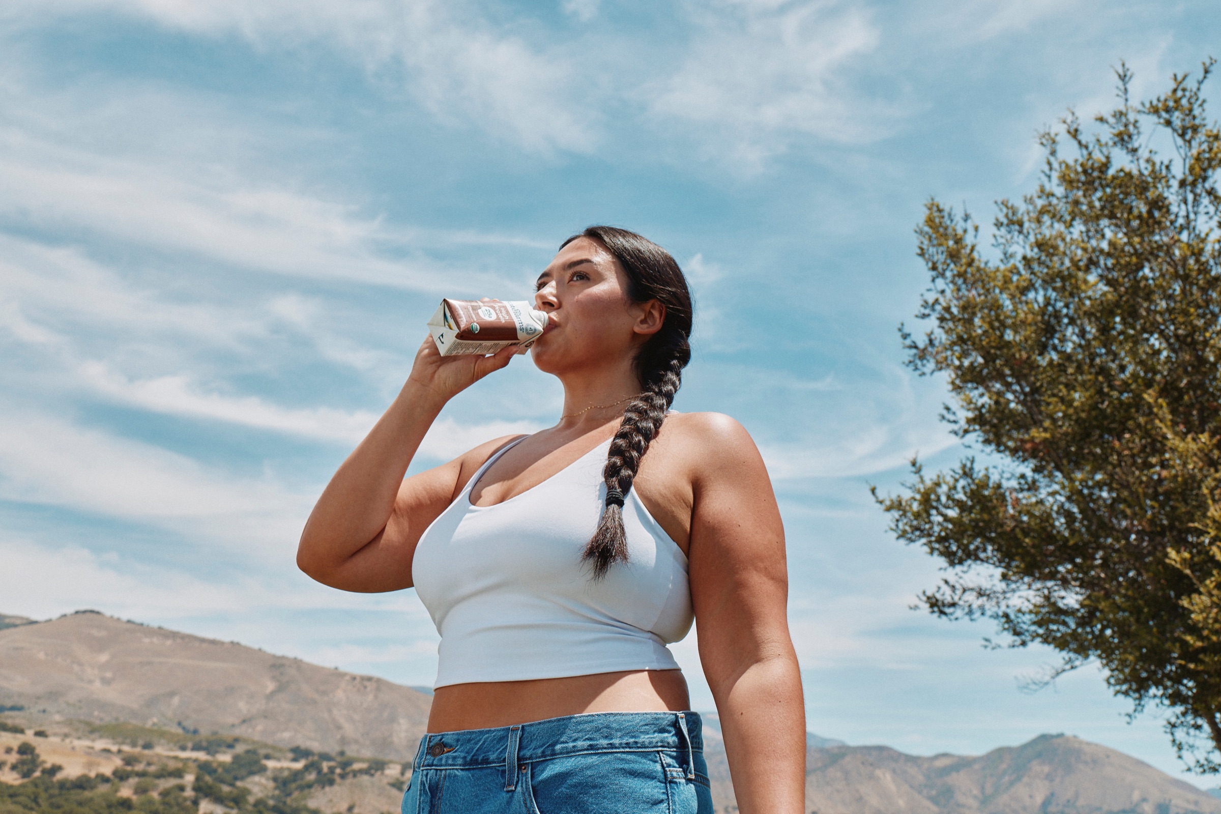 A woman drinking a Kate Farms nutrition shake outdoors against a blue sky and rolling hills.